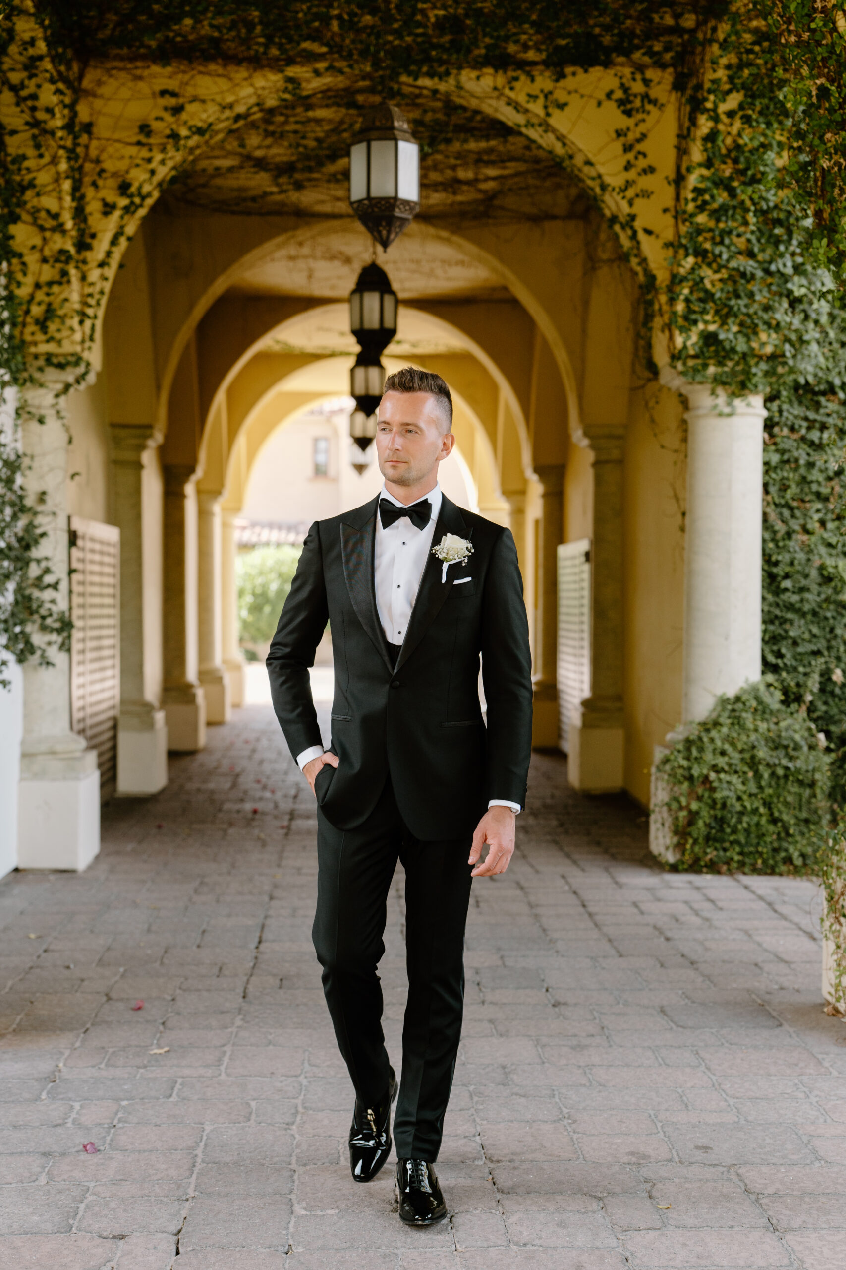 The groom walks confidently through an archway covered in ivy, his classic tuxedo perfectly tailored.