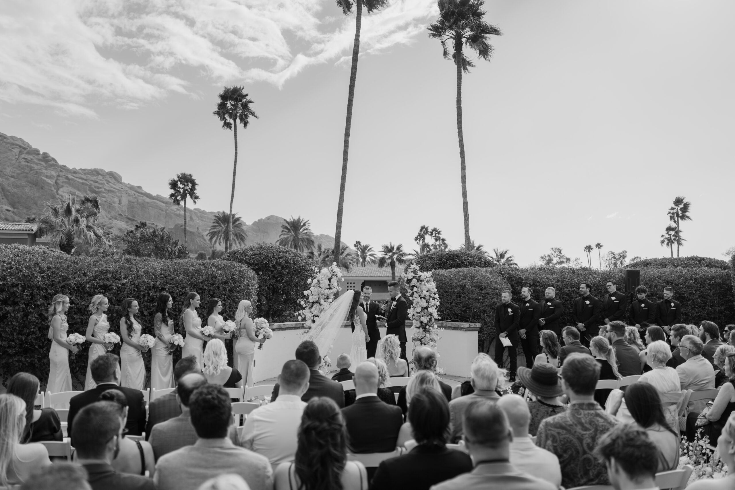 The couple exchanges vows at Omni Montelucia, framed by tall palm trees, floral arches, and a joyful crowd of loved ones.