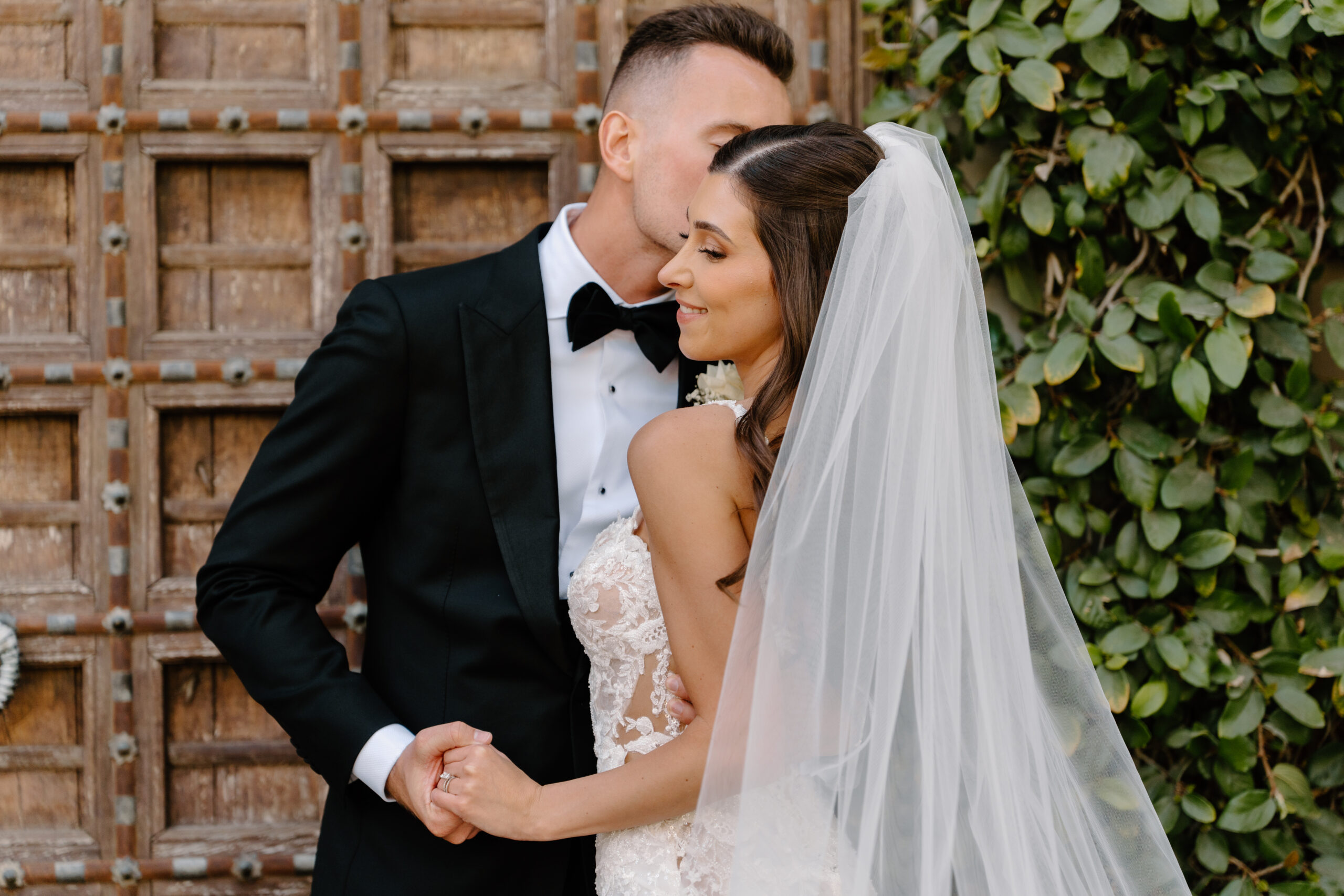 A romantic close-up of the groom kissing his bride’s temple as she smiles softly, framed by ivy and wooden doors.