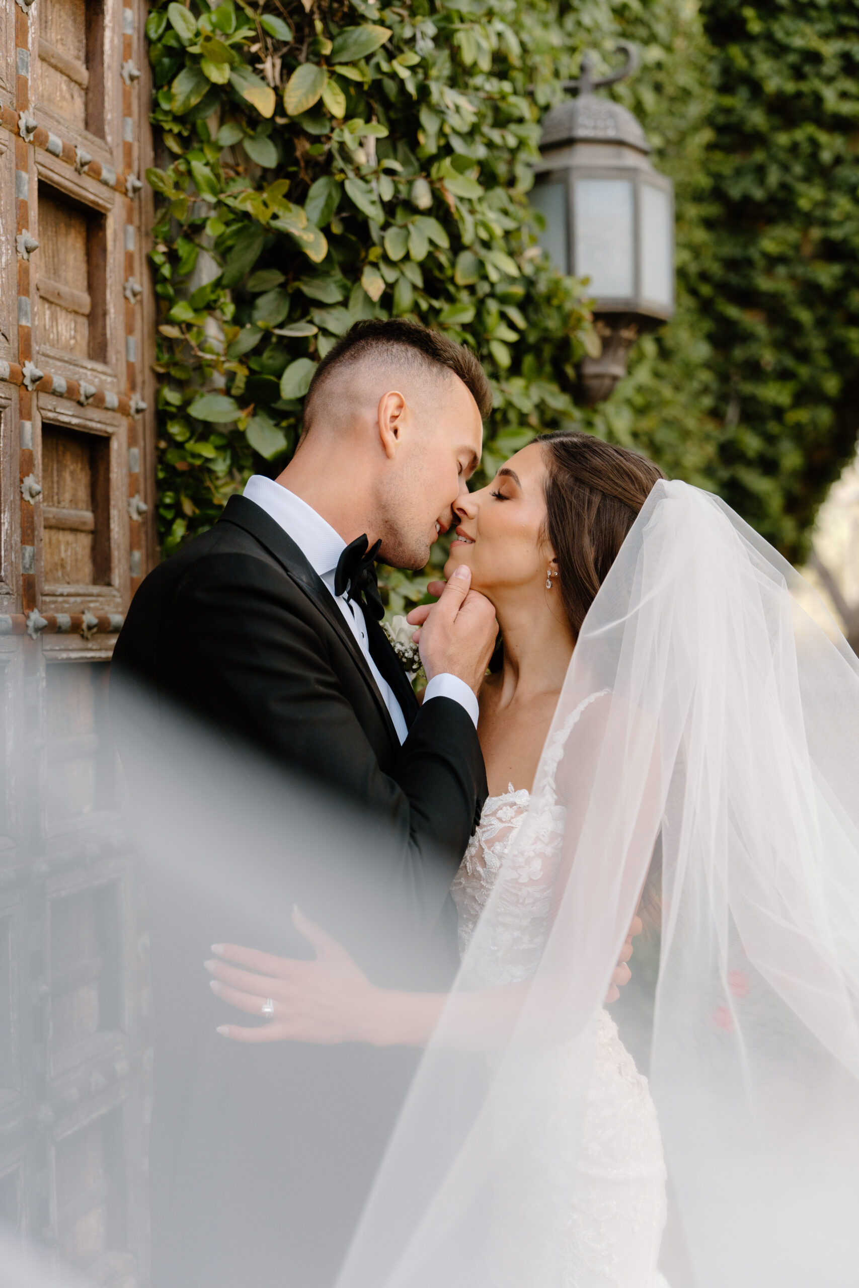 Bride and groom sharing a soft, intimate kiss beside an ivy-covered wall with her veil floating around them.