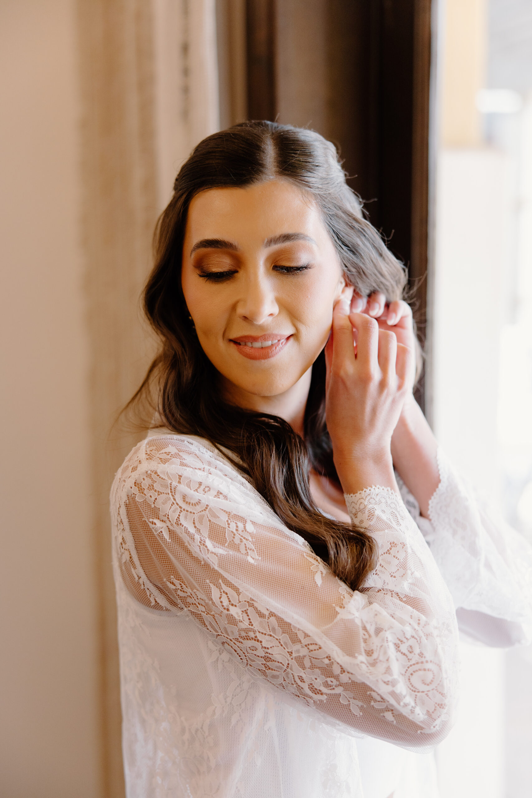 The bride adjusts her earrings by a window, the soft morning light highlighting her natural bridal makeup.
