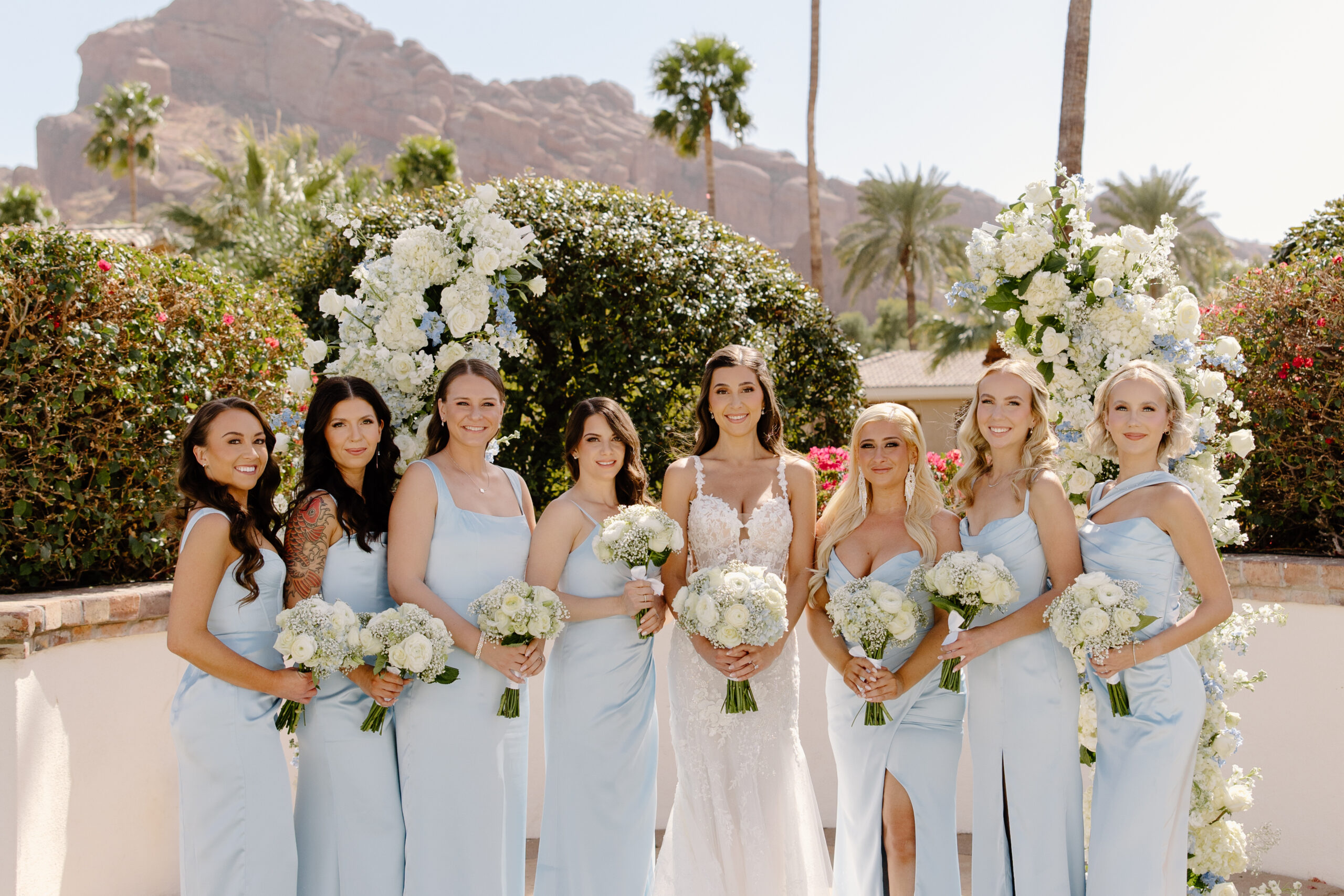Bride and bridesmaids posing together in soft blue dresses surrounded by lush blooms at the Omni Montelucia, with Camelback Mountain in the background.