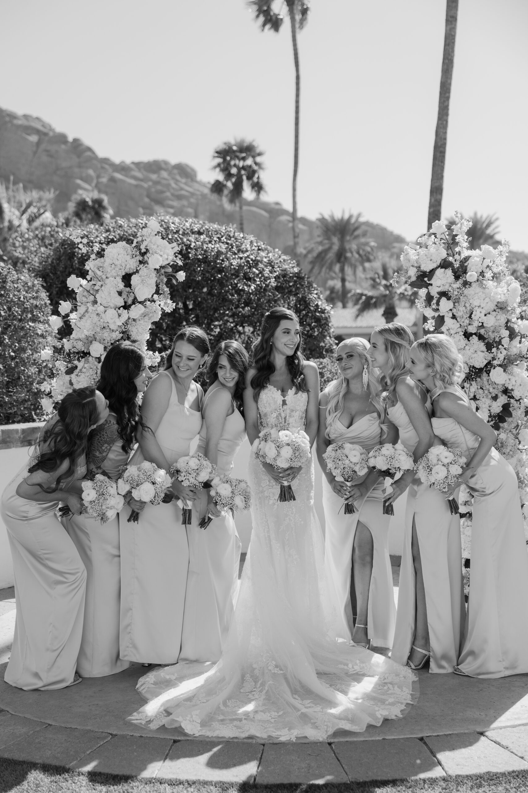 Bride smiling as her bridesmaids lean in playfully while holding white and blue bouquets.
