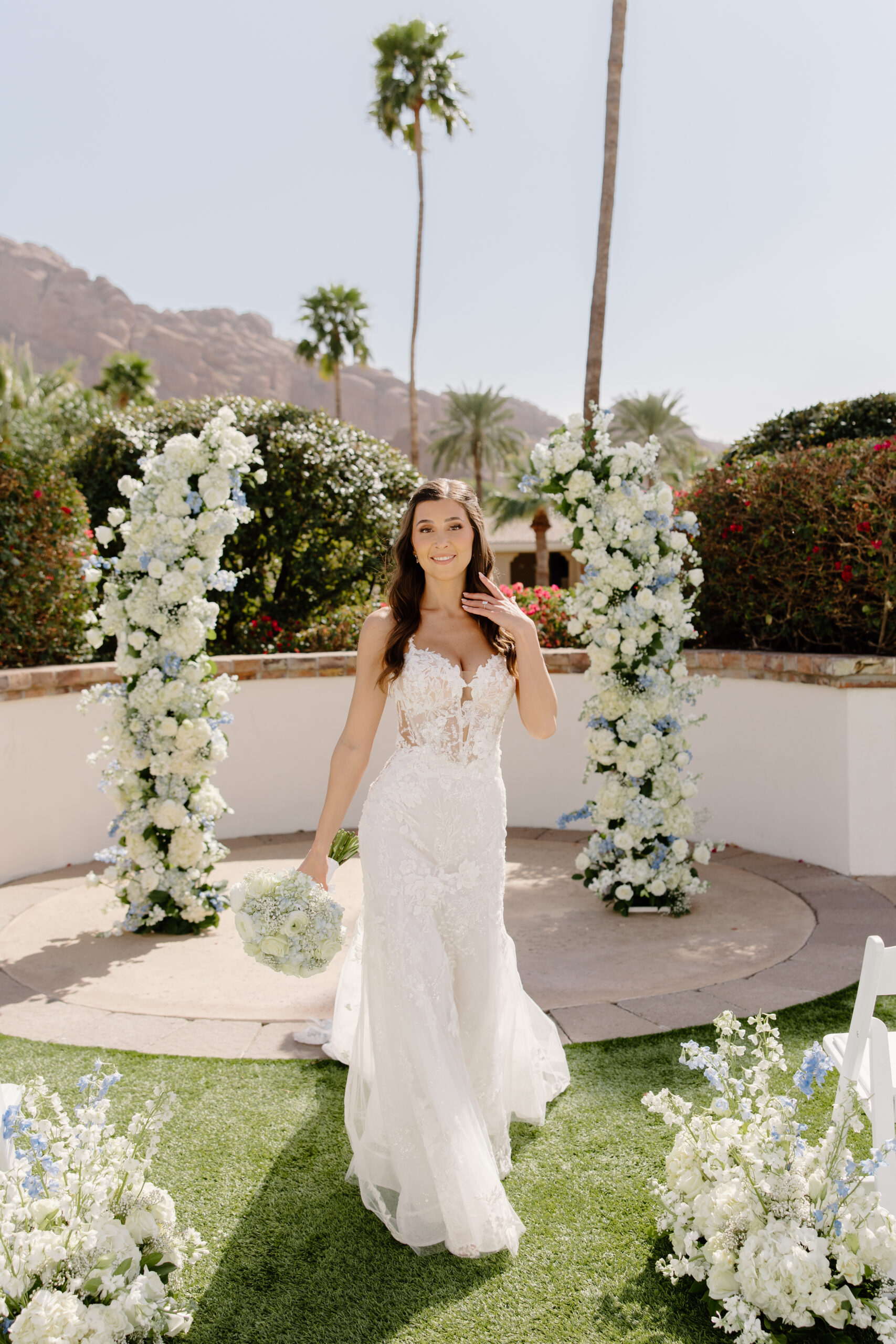 Bride walking down a flower-lined aisle at the Omni Montelucia, holding a white bouquet with Camelback Mountain and palm trees in the background.