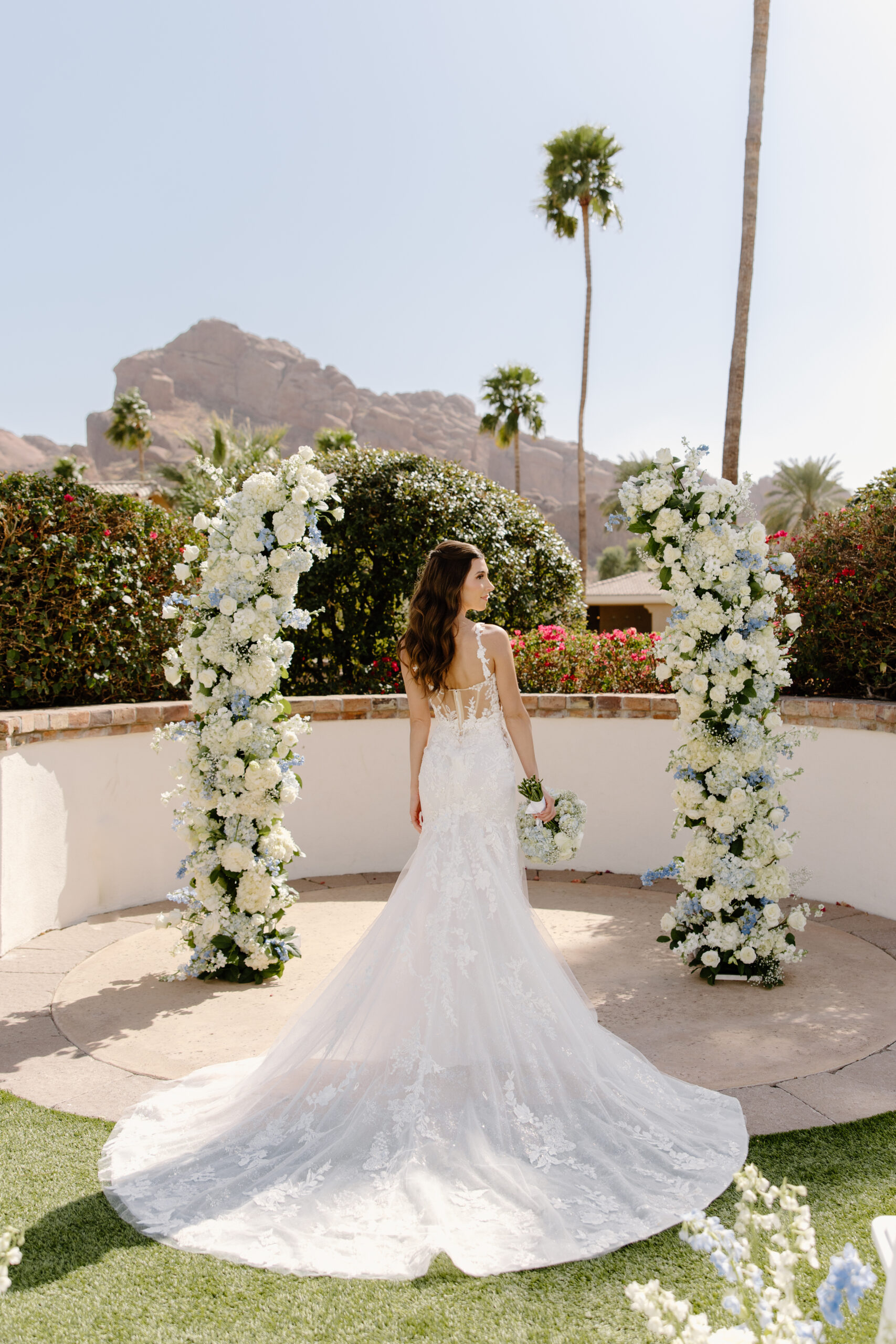 The bride stands between two floral pillars, her train cascading behind her with mountain views in the distance.