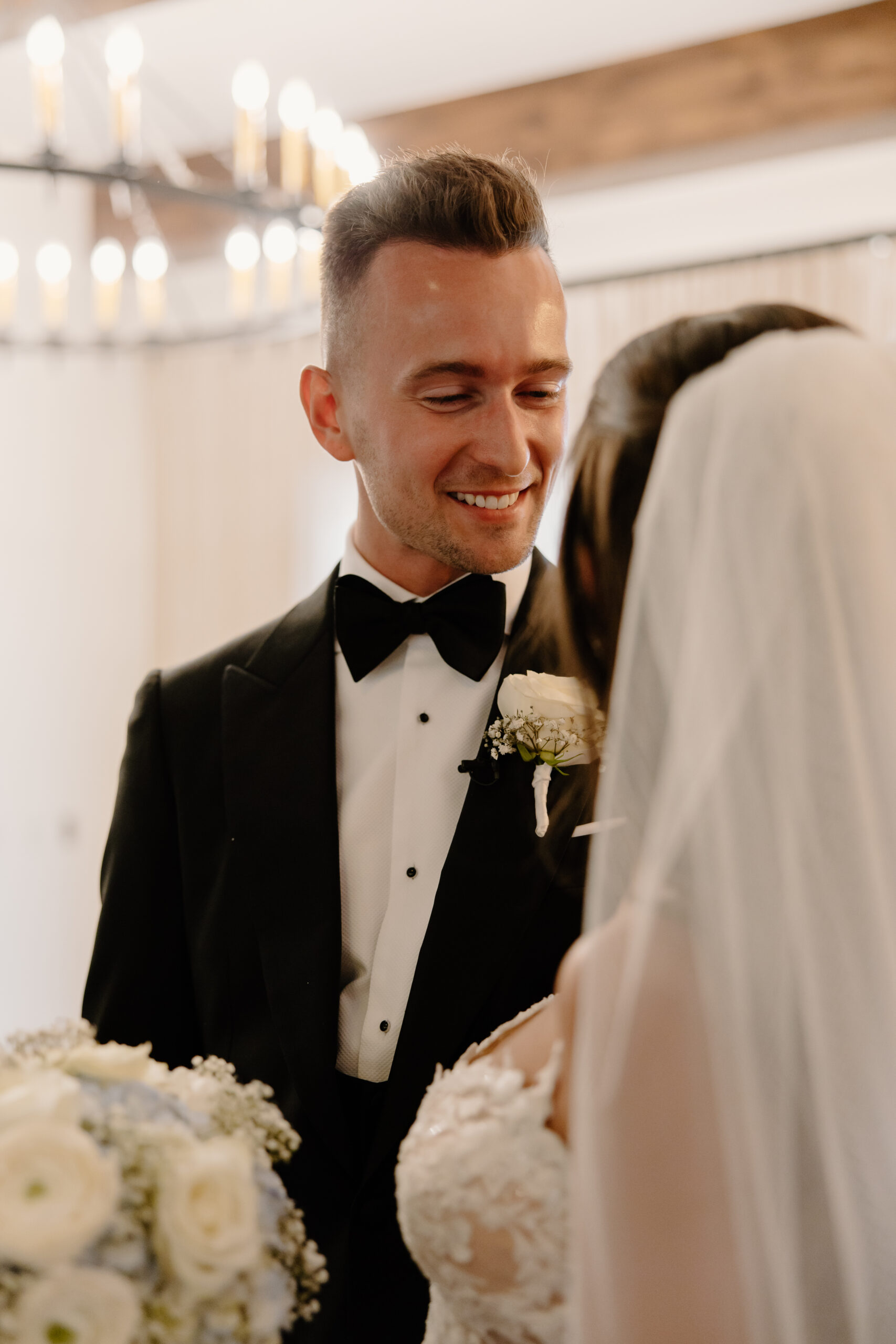 The groom smiles softly at his bride during their first look, holding her bouquet of white and blue florals.