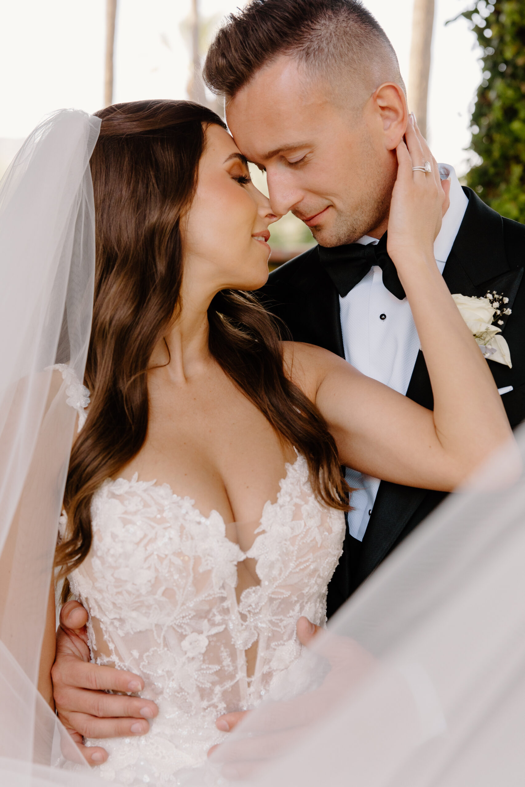The bride and groom walk hand in hand through a garden walkway, smiling toward each other as her veil trails behind.