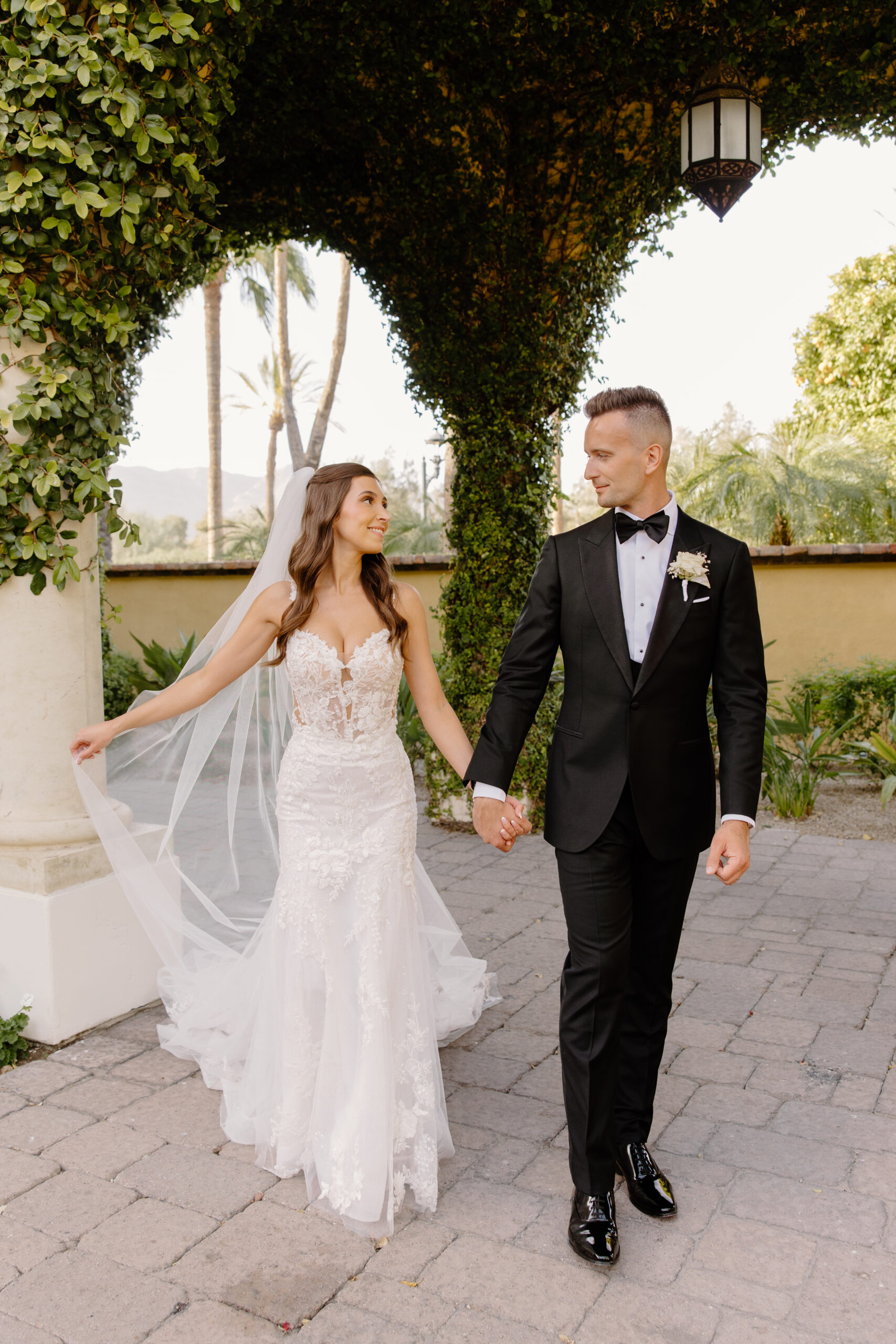 The bride and groom stand beneath the grand entry doors, her floral lace train spread beautifully across the terracotta floor.