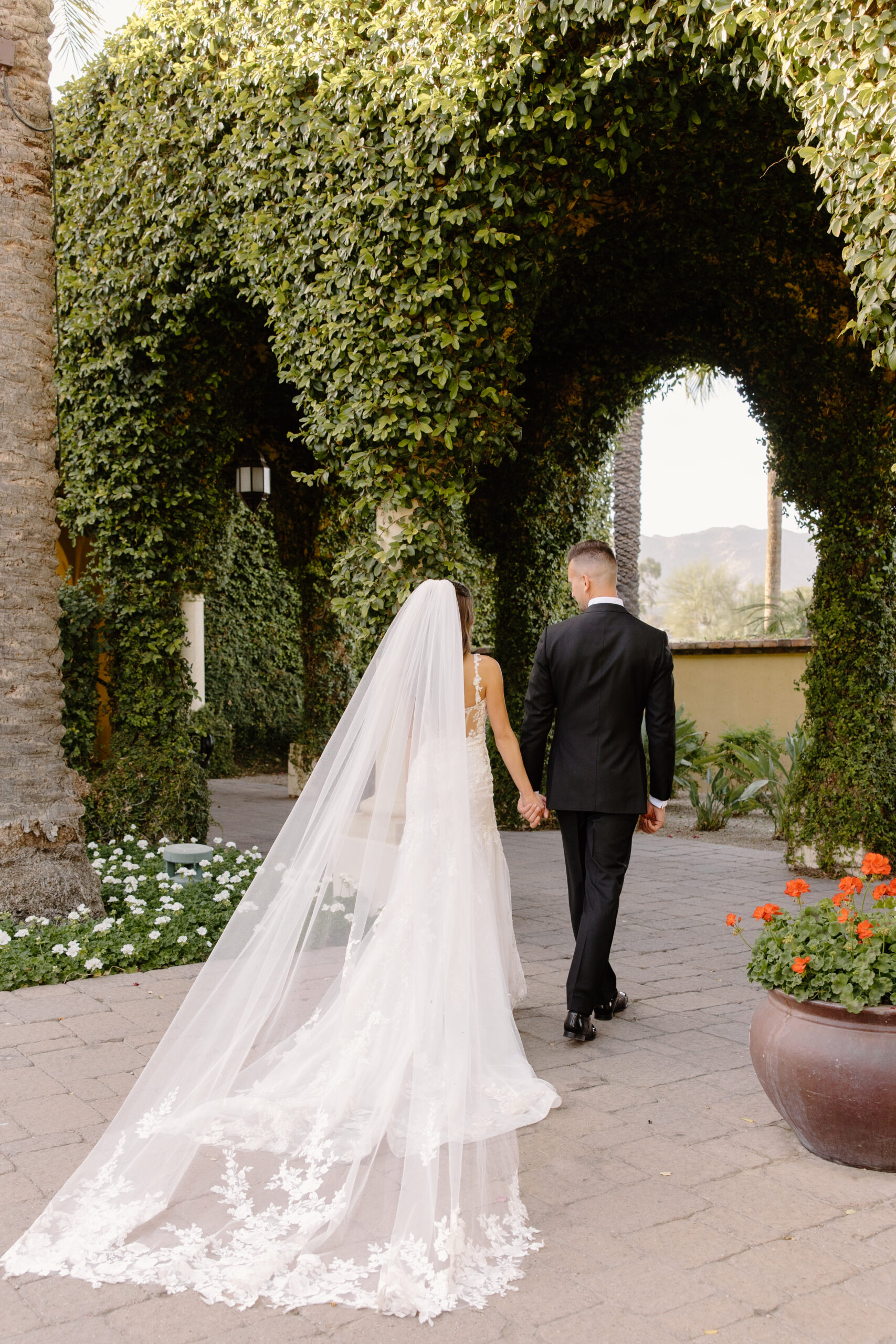 A romantic walk through Omni Montelucia’s courtyard, with the bride’s long veil trailing behind as they hold hands beneath green arches.