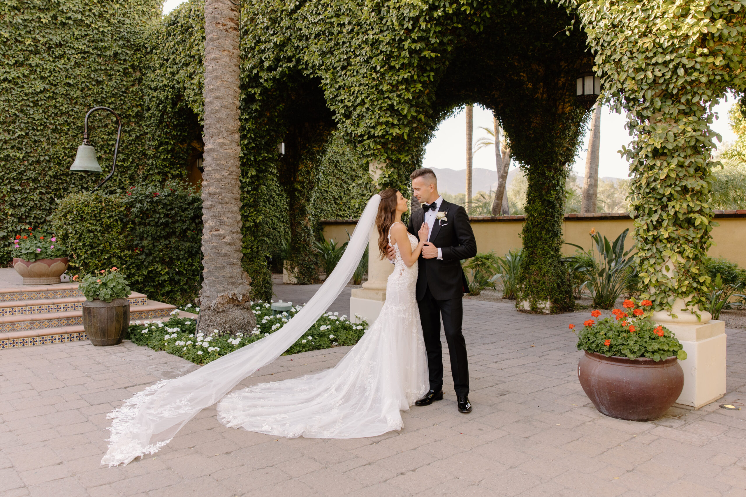The couple stands together beneath a canopy of greenery, the bride’s long veil trailing behind her in the quiet courtyard at the Omni Montelucia.
