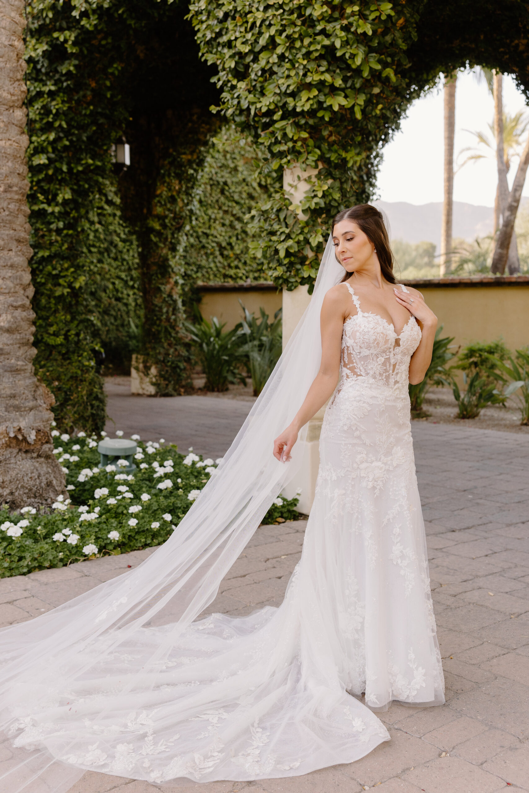 Bride posing gracefully under an ivy-covered arch, her long lace veil flowing behind her as she gently touches her dress.