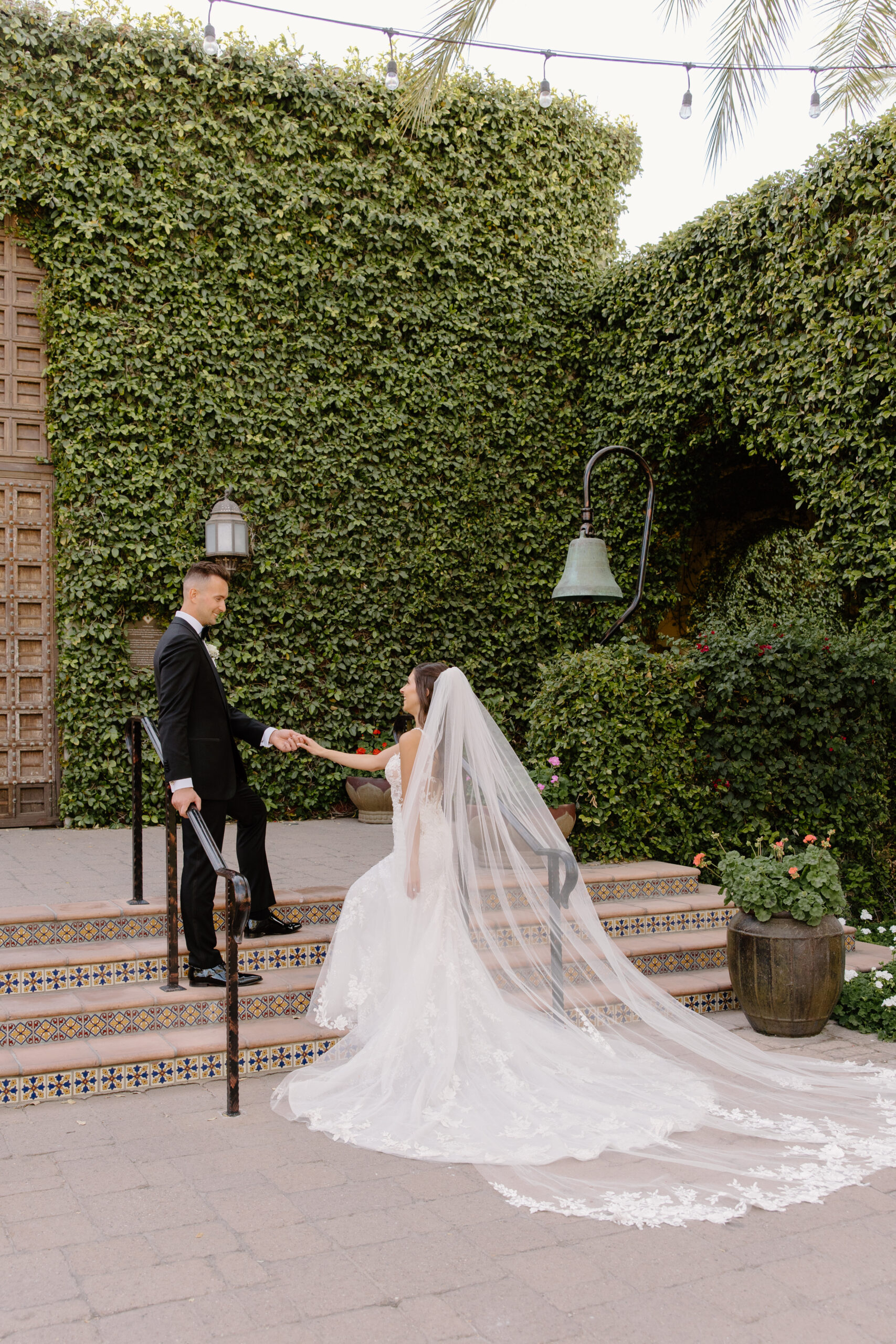 The groom gently holds his bride’s hand as she walks up tiled steps surrounded by lush greenery, her veil flowing behind her.