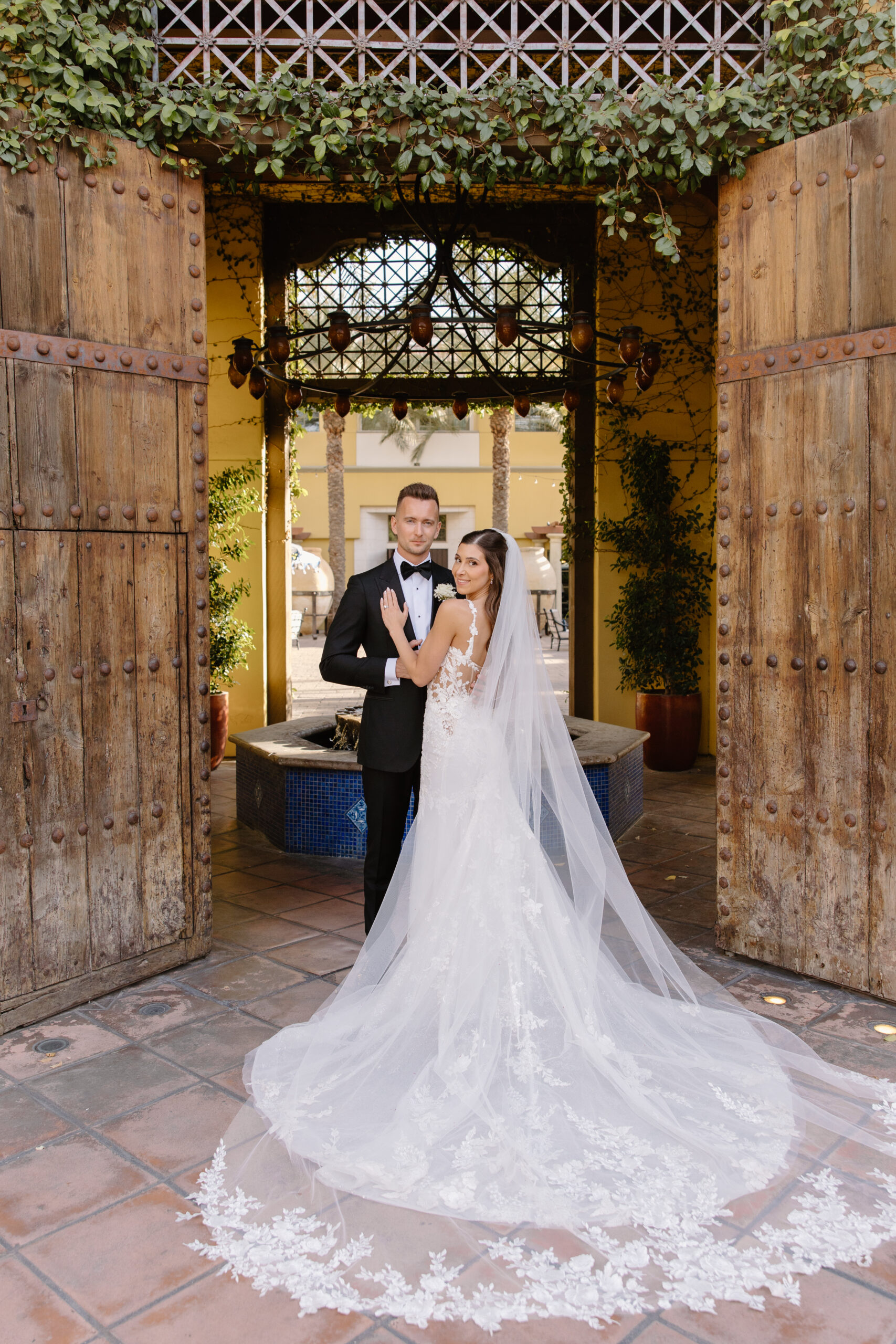 The bride and groom pose in front of rustic wooden doors, her lace gown flowing behind her on the tiled patio.
