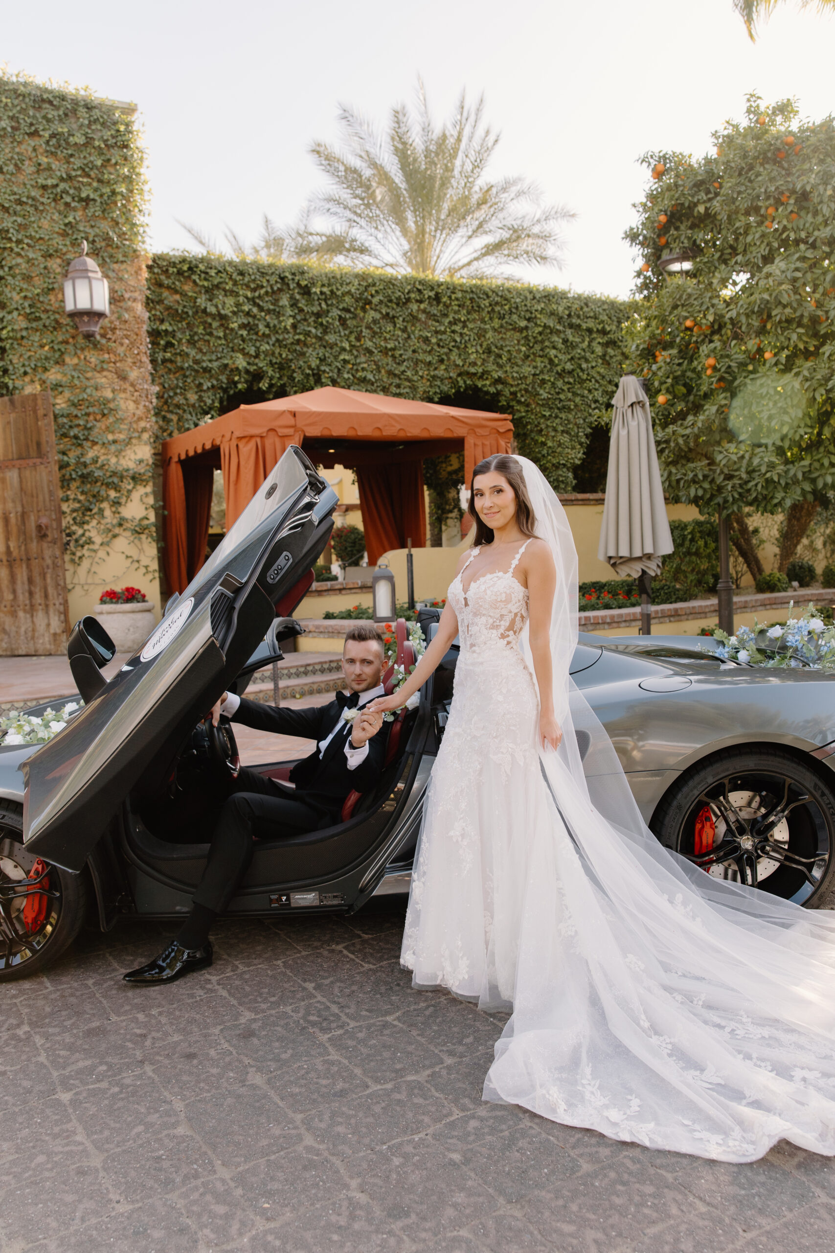 The bride and groom pose with their luxury car, her veil flowing behind her in the golden afternoon light.