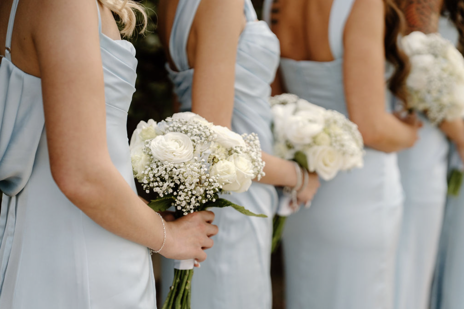 Bridesmaids in soft blue dresses holding white rose and baby’s breath bouquets.