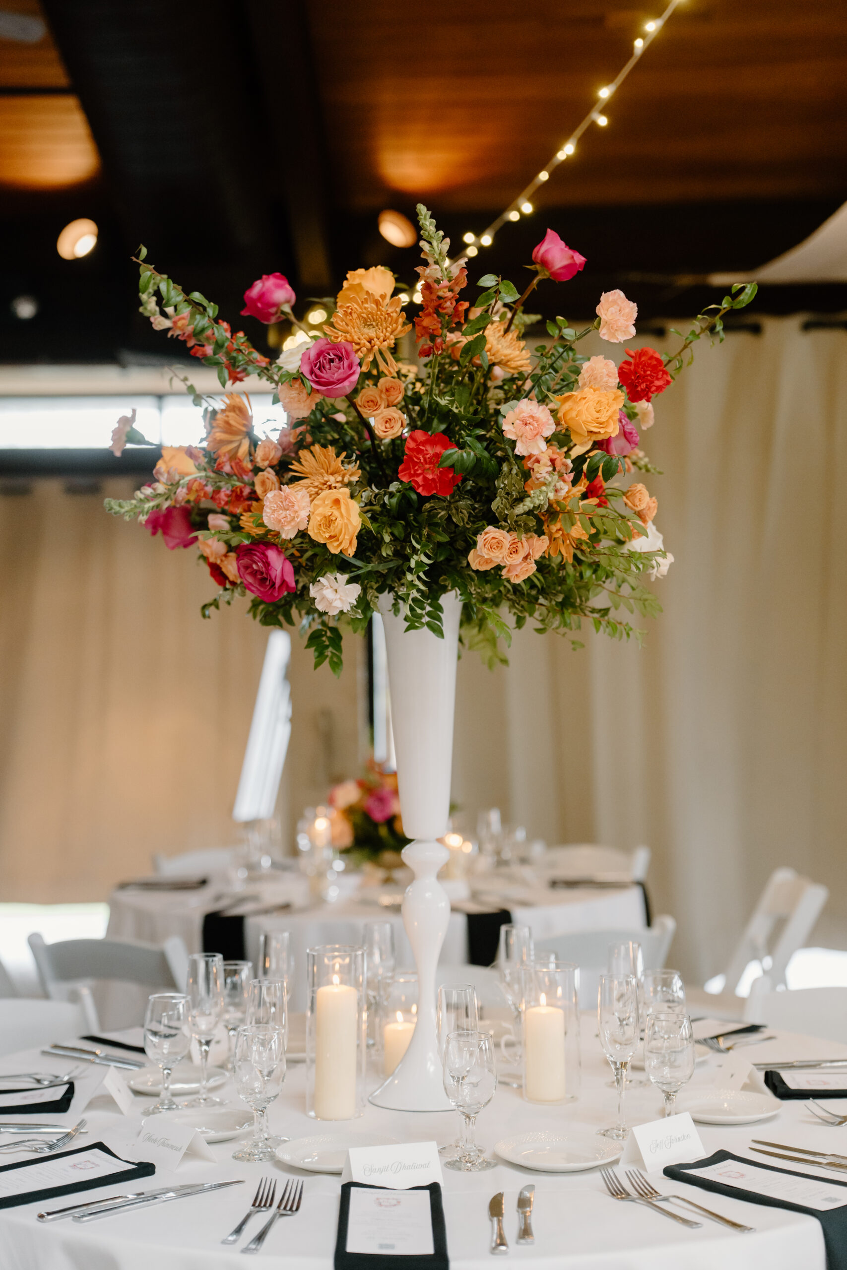 A tall centerpiece of colorful roses and greenery on a reception table surrounded by candles and elegant table settings.