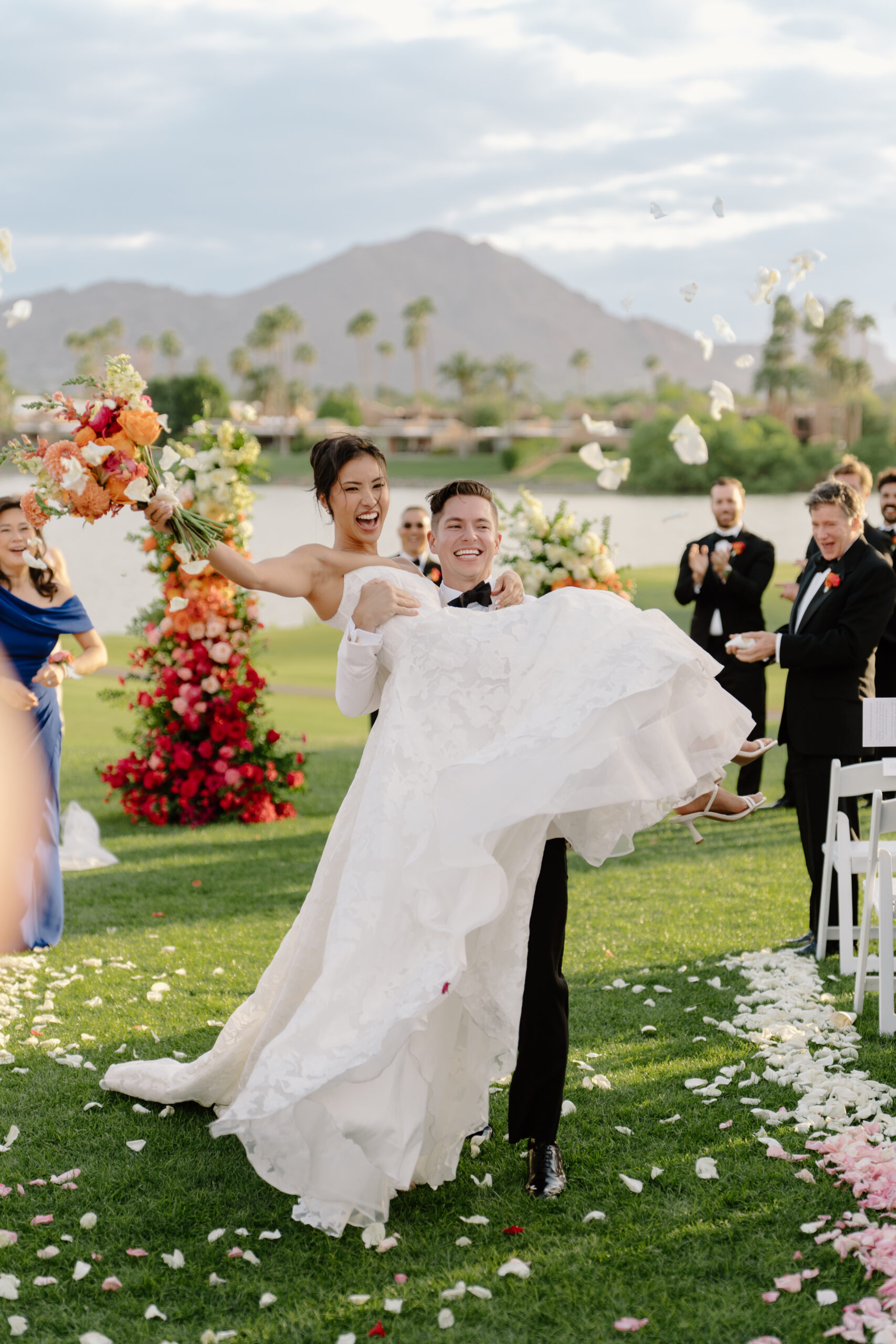 The groom joyfully lifting the bride as guests cheer and flower petals fall around them during their ceremony at mccormick ranch golf club.
