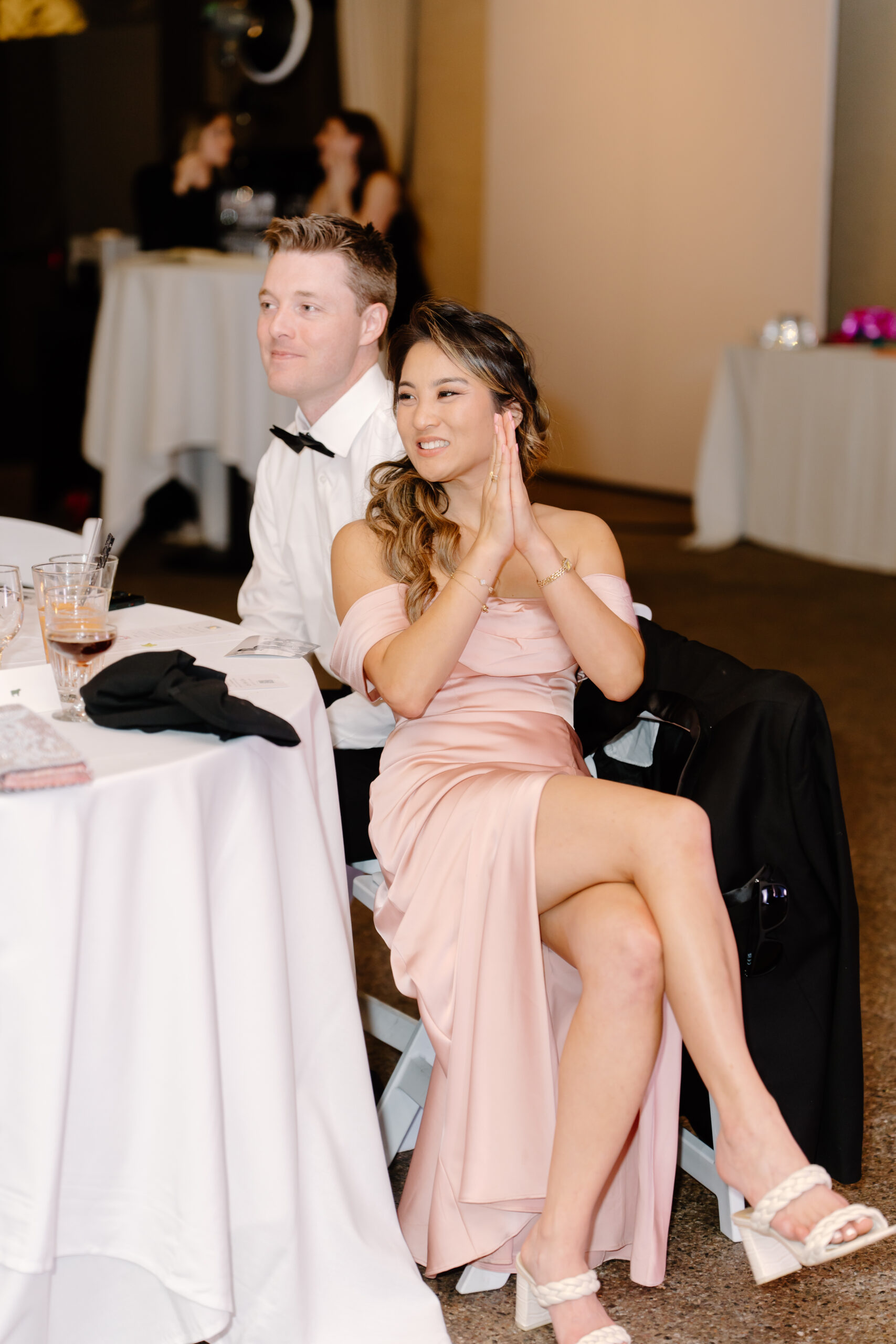 Bridesmaid seated at a table smiling and clapping while watching speeches during the reception.