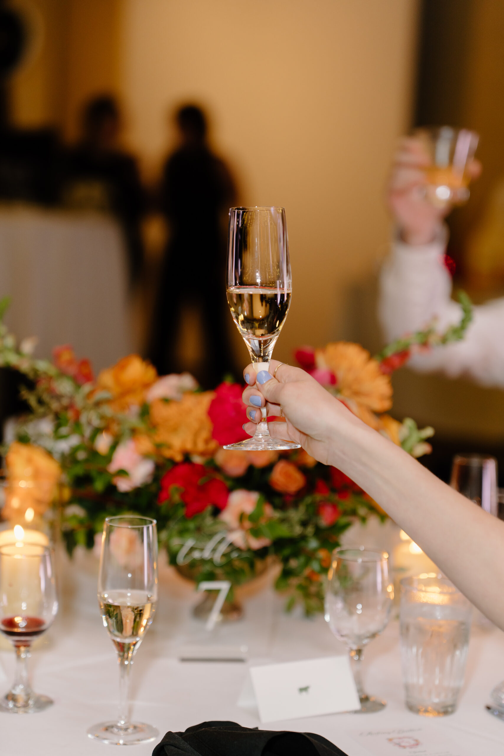 A hand raising a champagne glass during the reception, with colorful floral centerpieces and candlelight on the table.