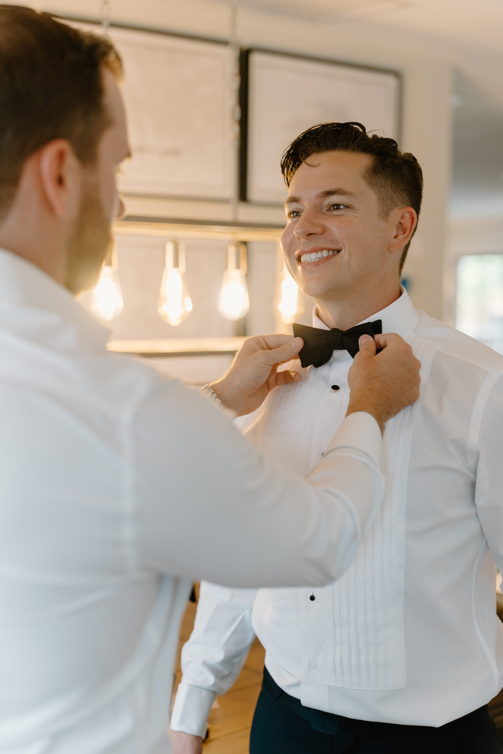 A groomsman helping adjust the groom’s bow tie while he smiles just before the ceremony.
