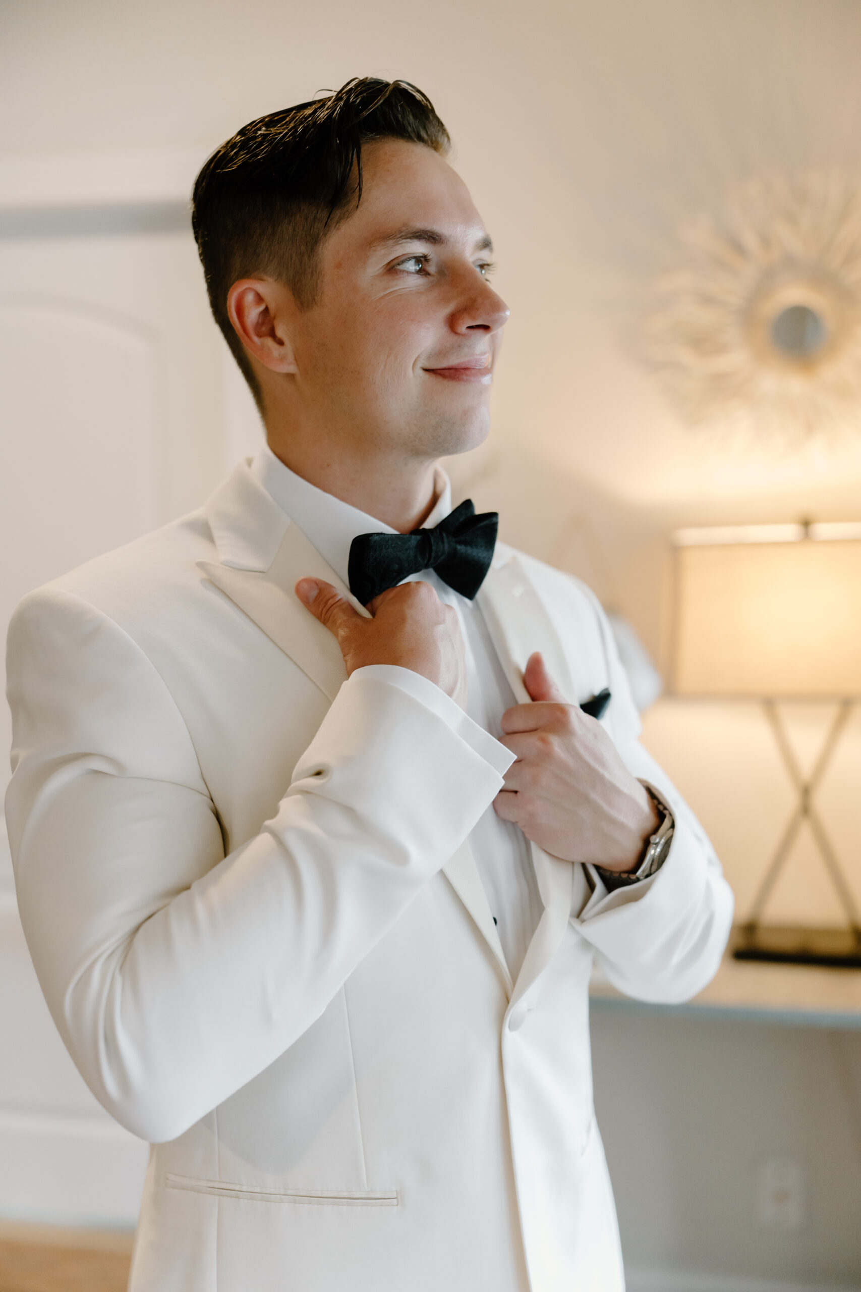 A groom adjusting his bow tie indoors, smiling softly as he finishes getting ready.