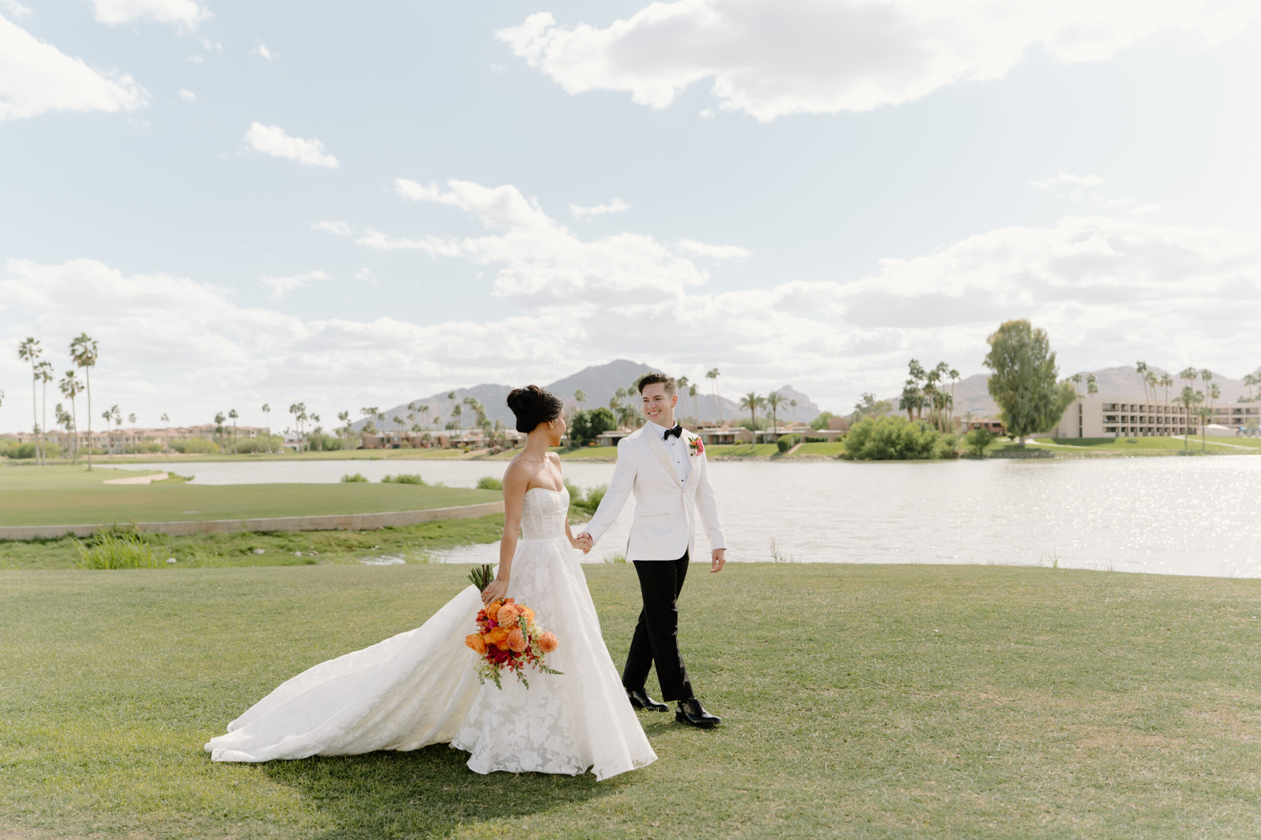 Bride and groom walking hand in hand with a colorful bouquet and mountain views reflected in the water behind them at McCormick Ranch Golf Club.