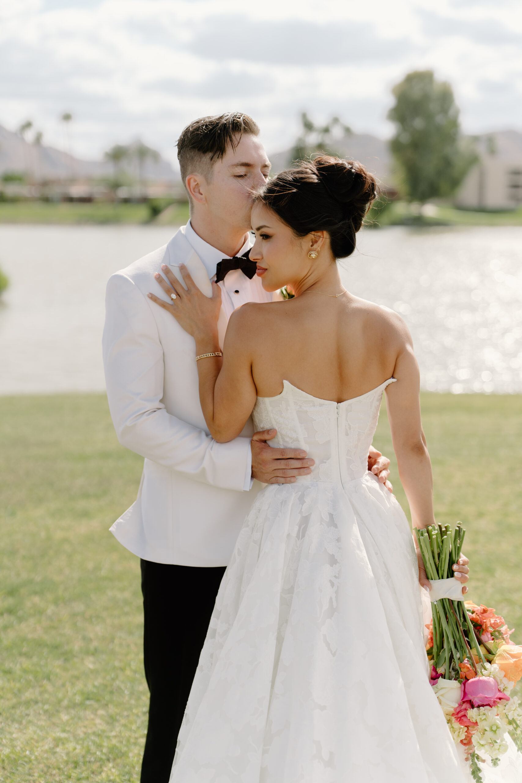 Groom kissing the bride’s temple as she holds her bouquet, standing lakeside beneath soft afternoon light at McCormick Ranch Golf Club.