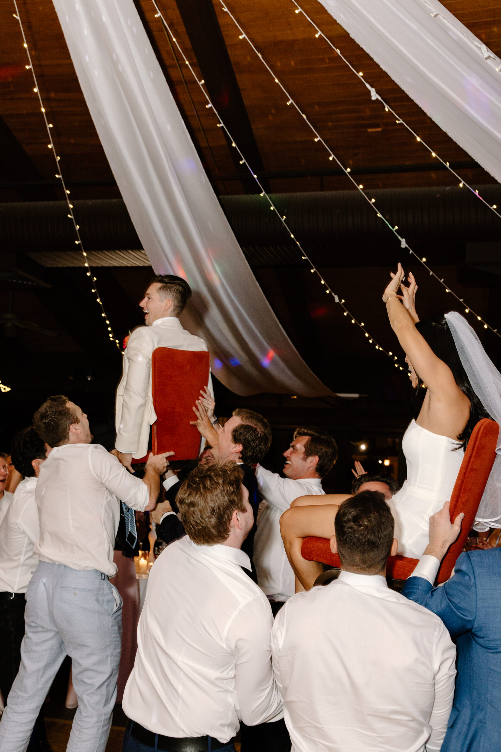 Guests lifting the bride and groom on chairs during the hora, cheering and dancing around them.