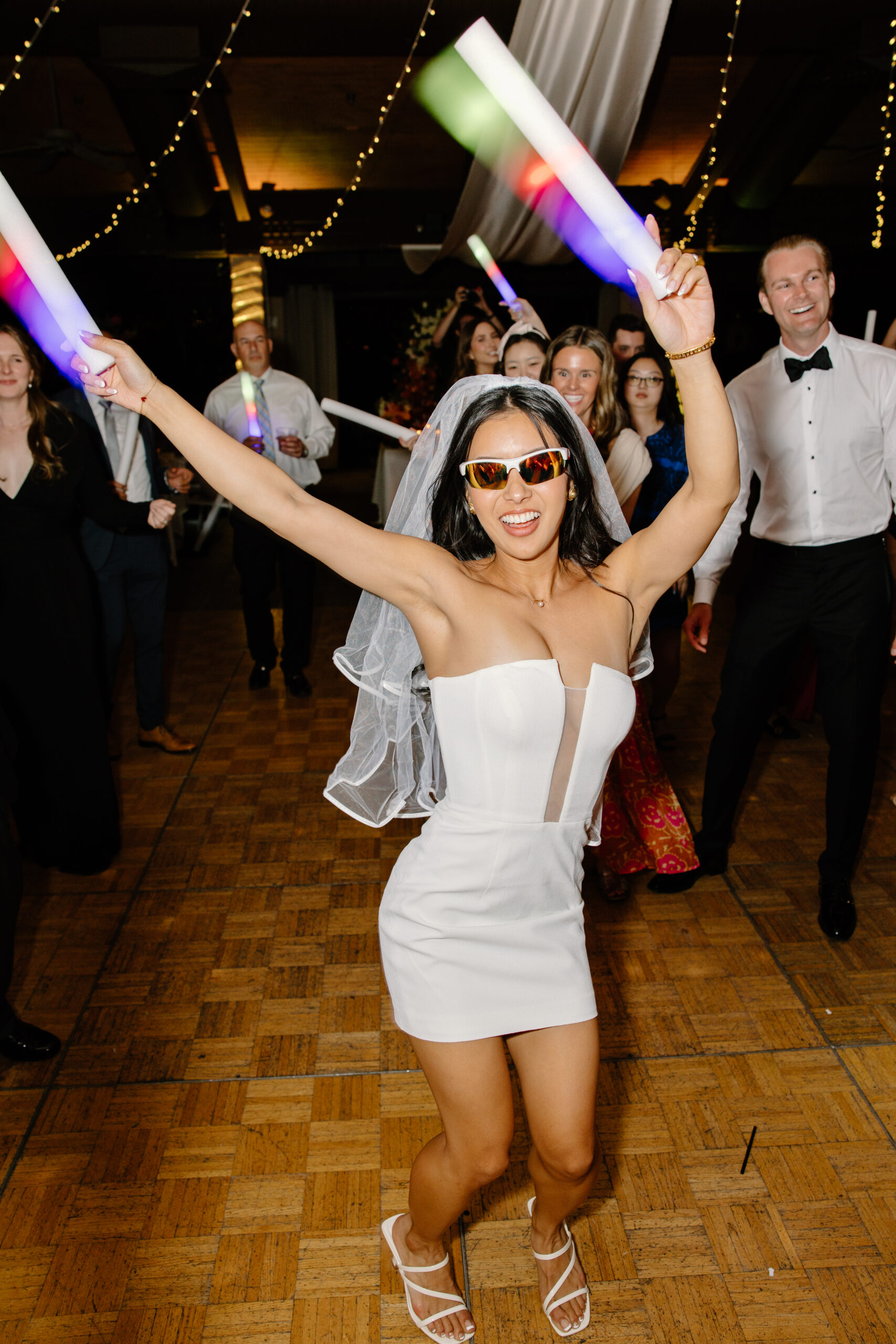 Bride wearing a short reception dress and sunglasses while dancing with glow sticks on the dance floor.