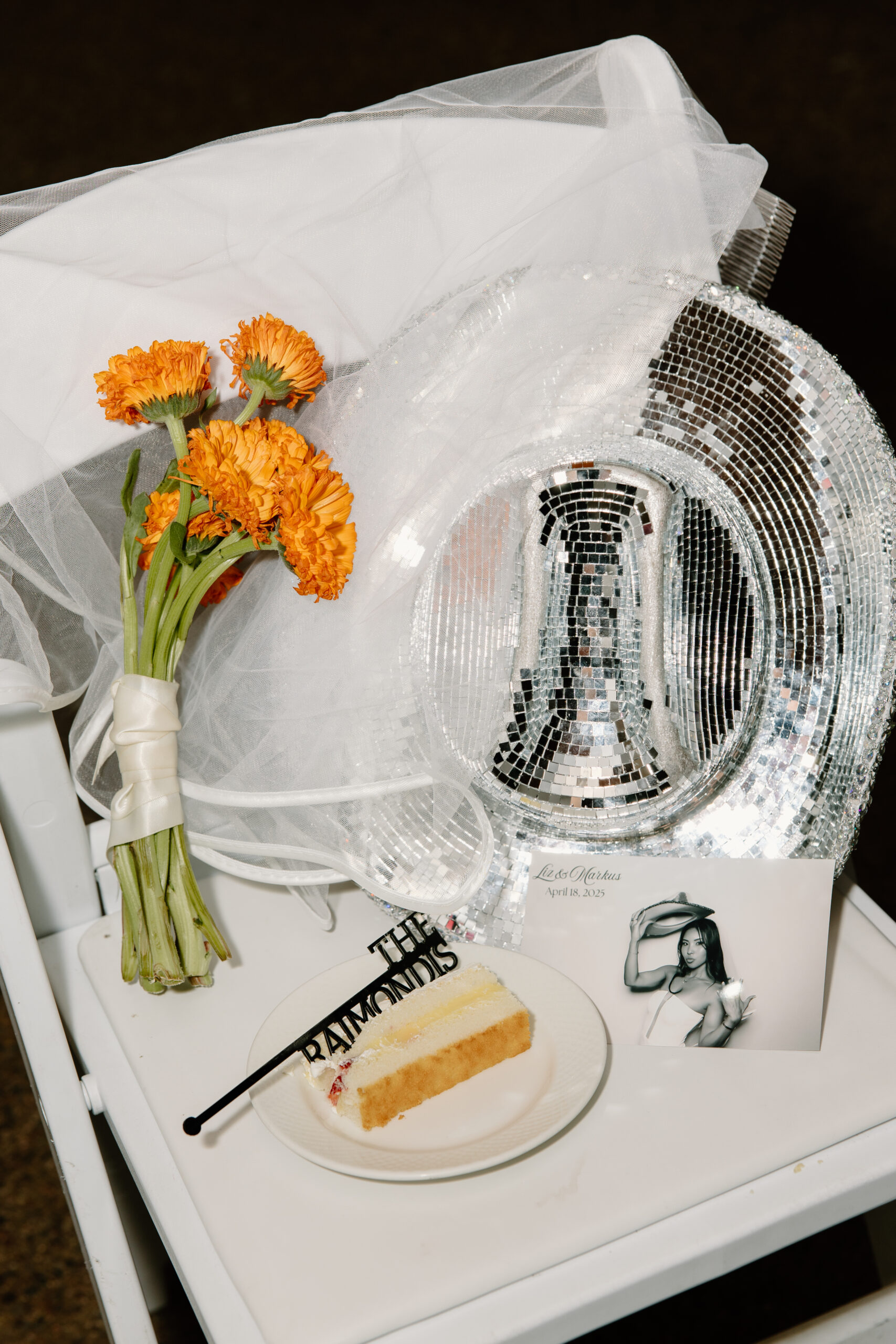 A styled flat lay featuring a disco-ball cowboy hat, orange flowers, a slice of cake, and a wedding photo displayed on a small table.