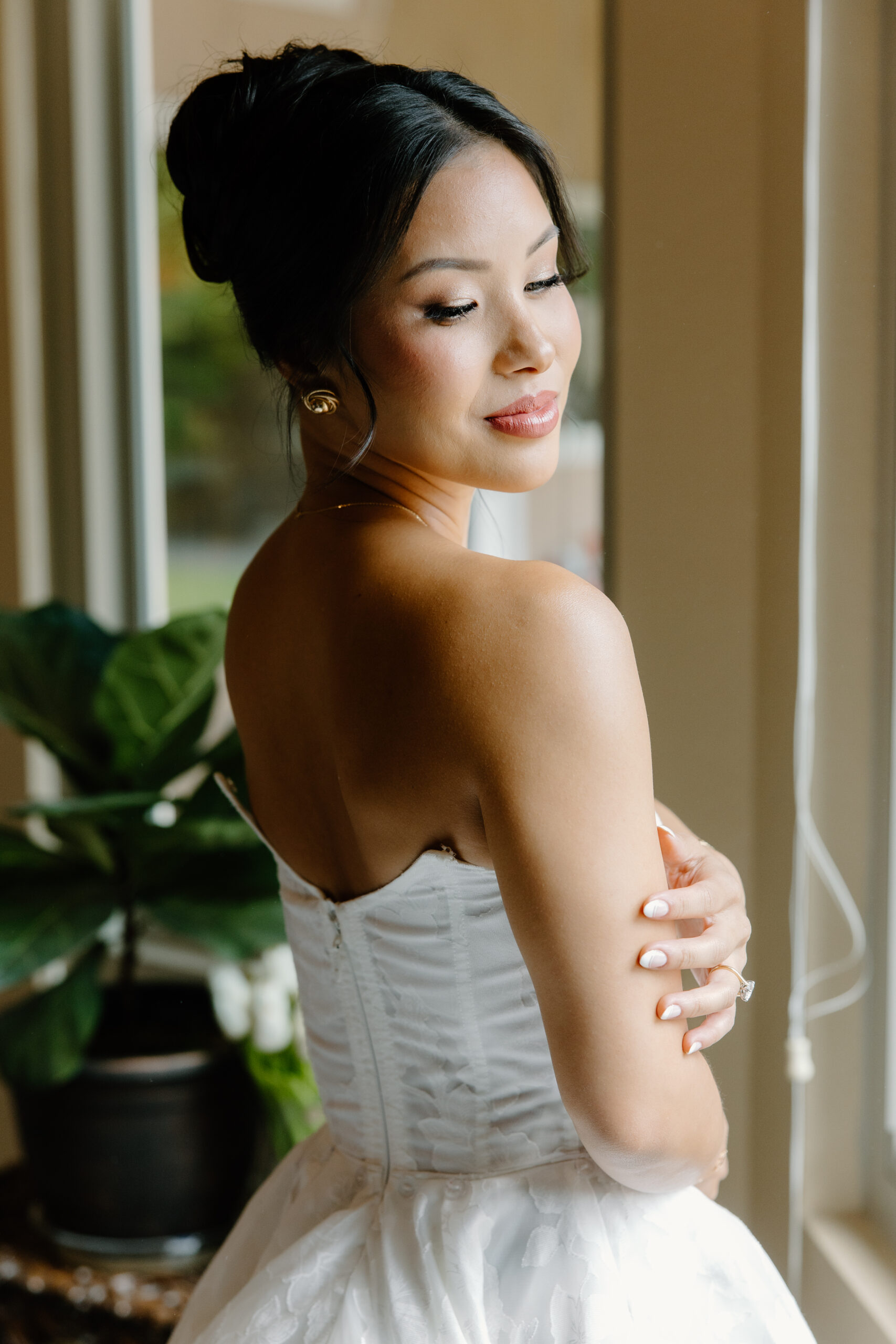 A close-up portrait of the bride near a window, gently touching her arm and smiling with her hair styled in an elegant updo.