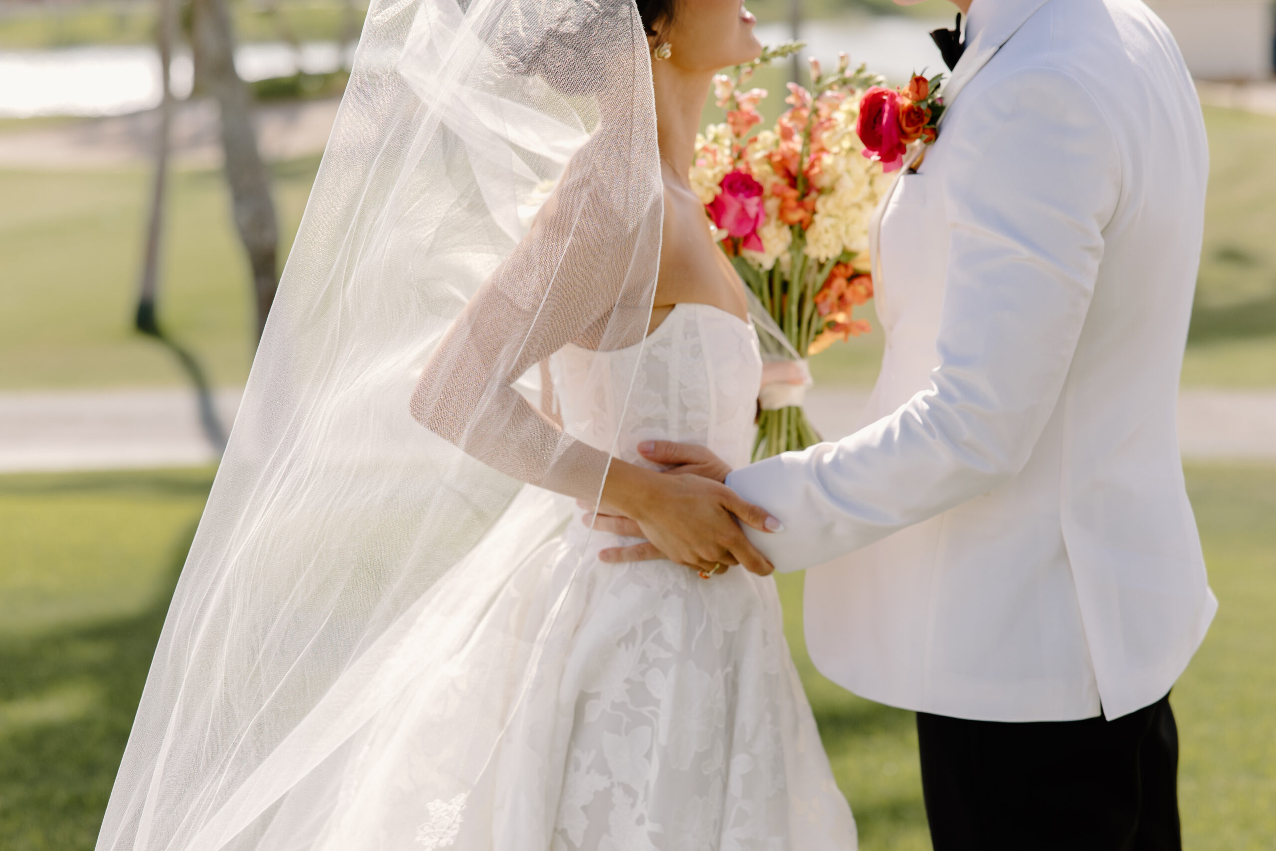 A close-up of the bride and groom holding each other, her veil blowing gently as she holds a bright bouquet of coral and peach flowers at mccormick ranch golf club.