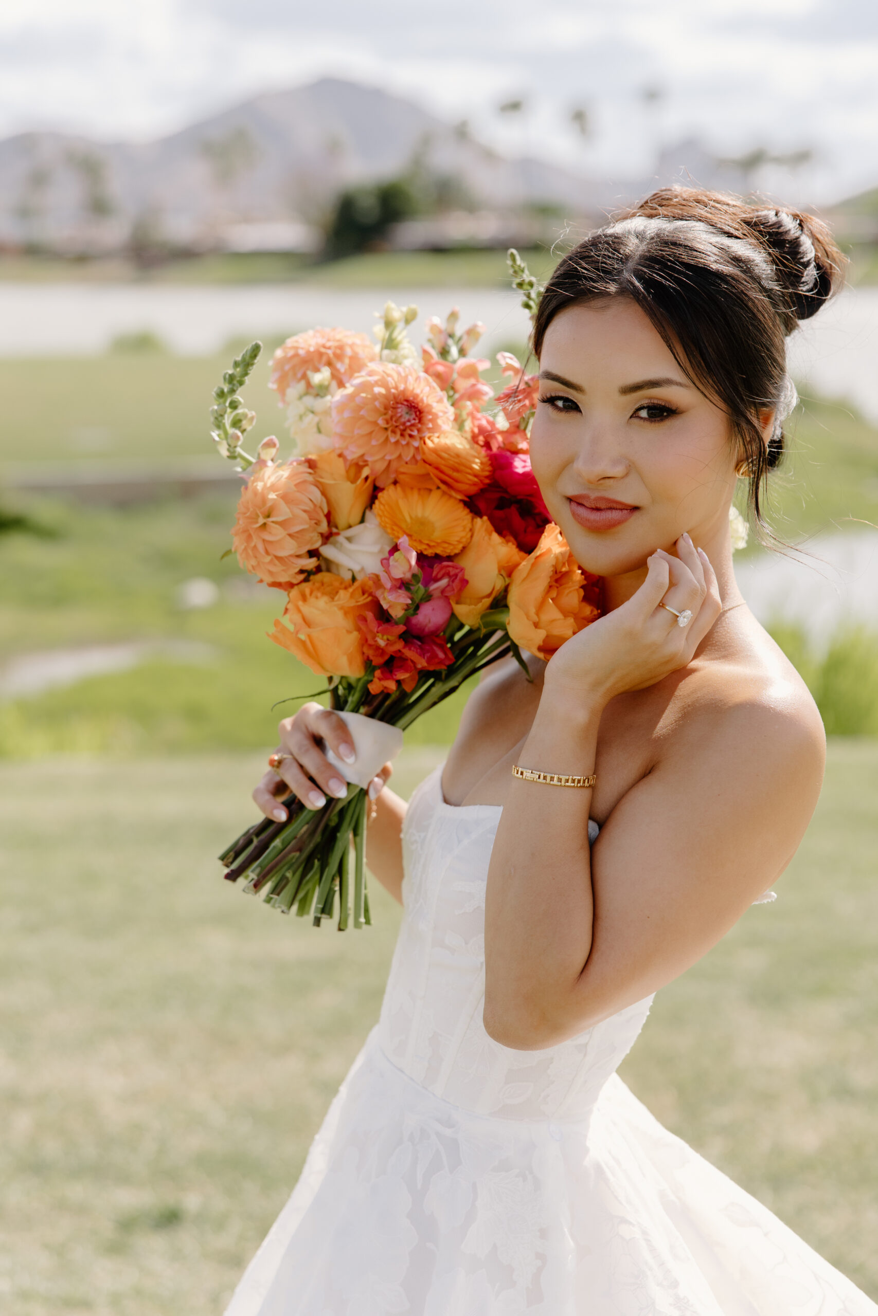 A bride holding a vibrant bouquet of orange and pink flowers while smiling softly toward the camera.