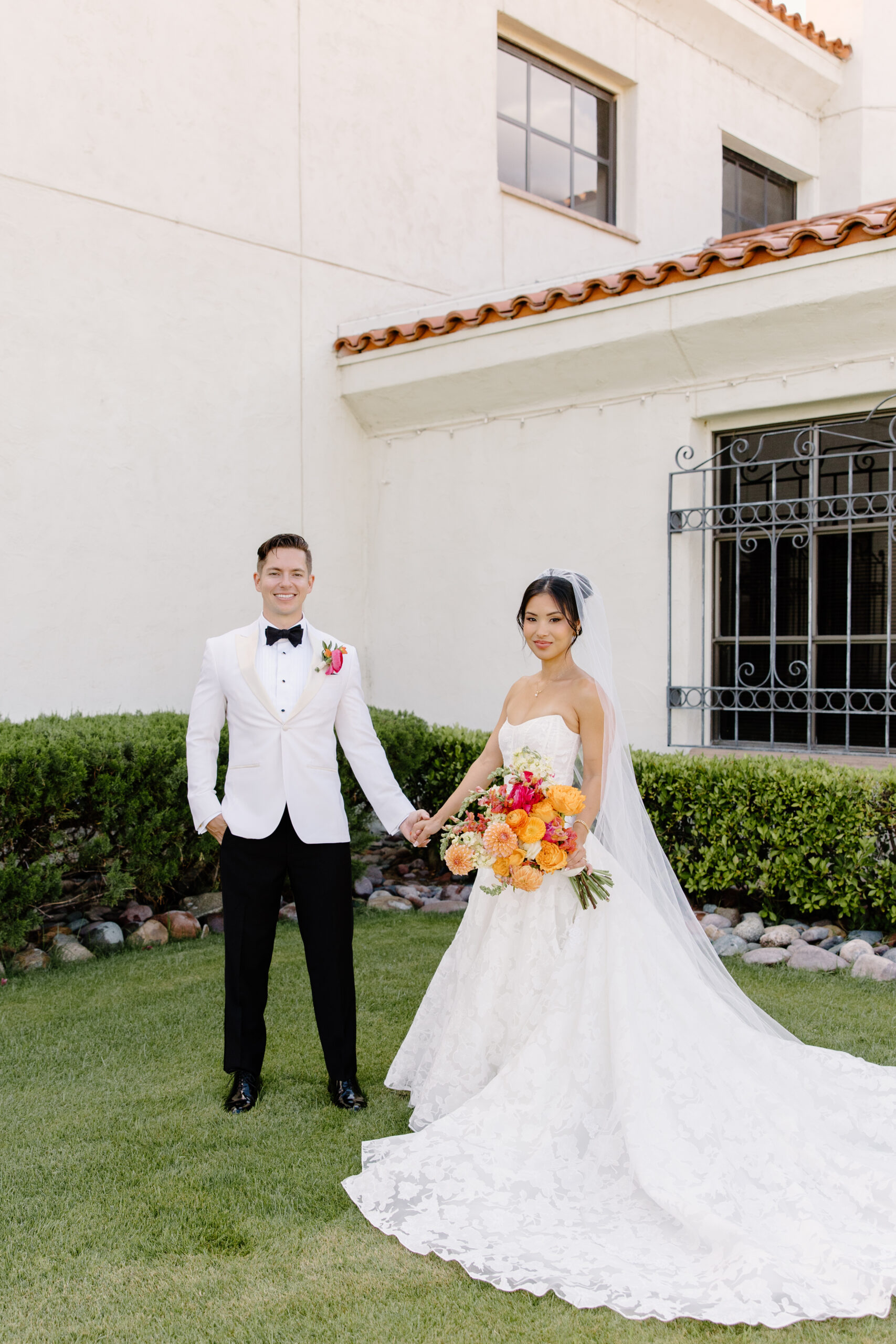 The couple standing hand-in-hand against the cream-colored exterior of the venue, her lace train spread across the grass at mccormick ranch golf club.