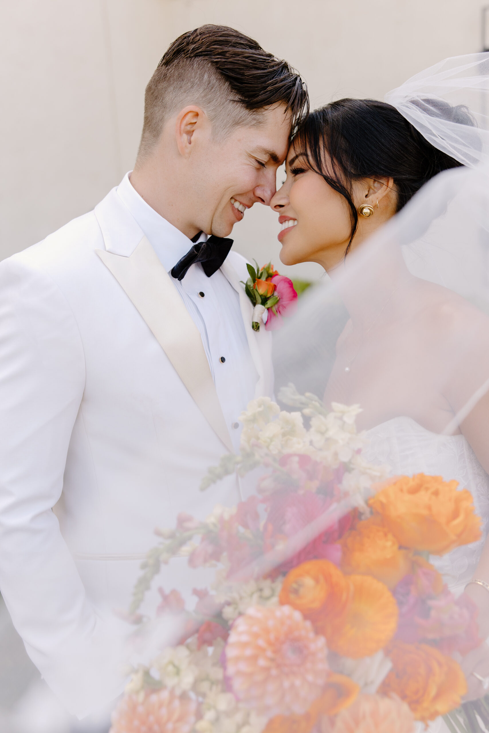 A close, intimate moment as the bride and groom lean forehead-to-forehead, her bouquet of orange and pink flowers in the foreground.