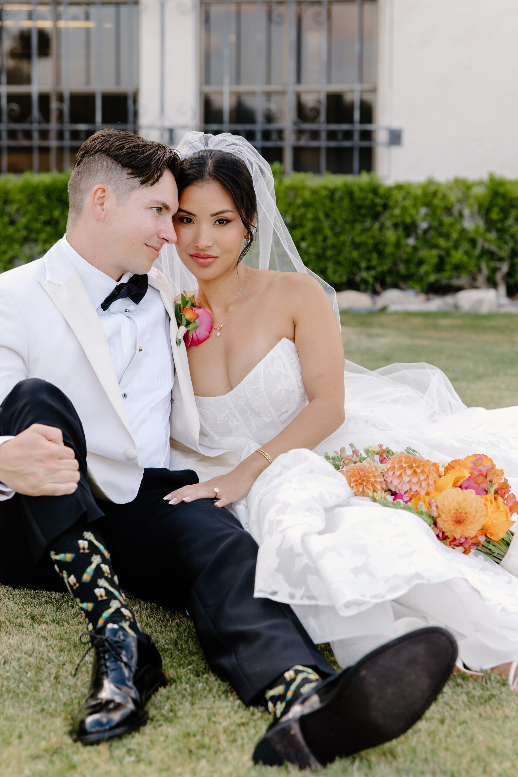 Bride and groom sitting together on the lawn, holding each other closely with bright florals resting beside them.