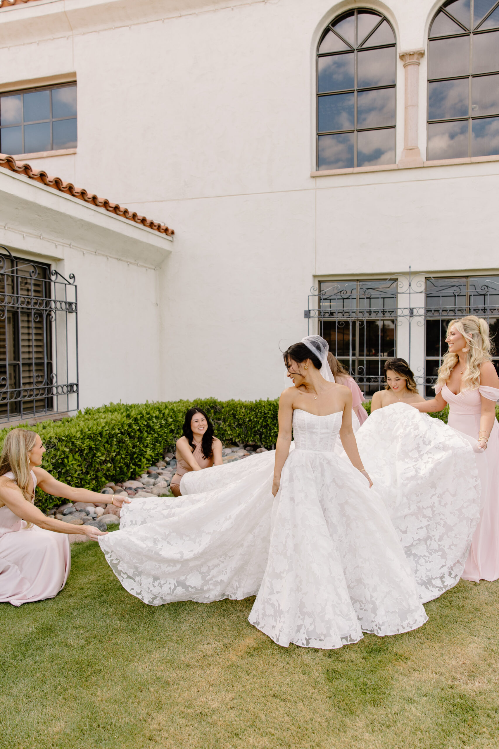 Bridesmaids helping adjust the bride’s full lace gown train outside a white Mediterranean-style building.