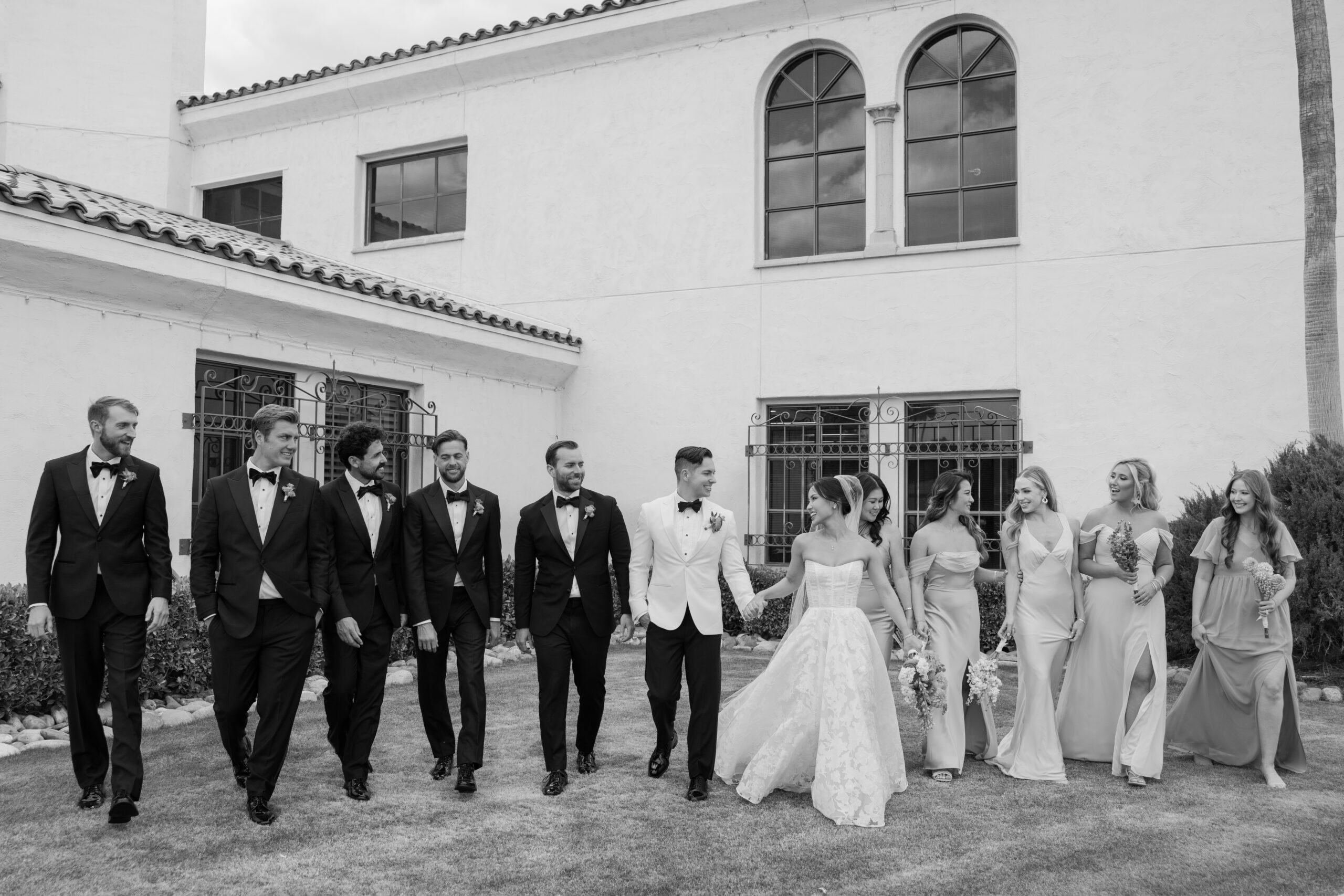 Black and white photo of the wedding party walking together outdoors, laughing and smiling as they line up beside the couple.