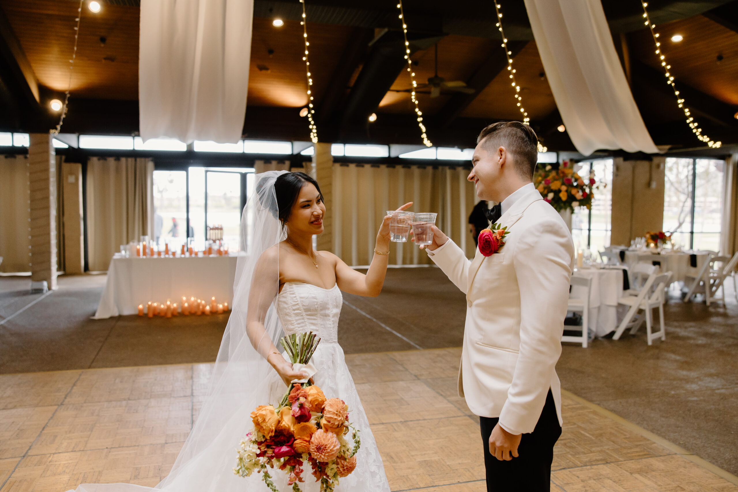 The couple toasting each other inside the reception space at mccormick ranch golf club, surrounded by twinkle lights and draped fabric.