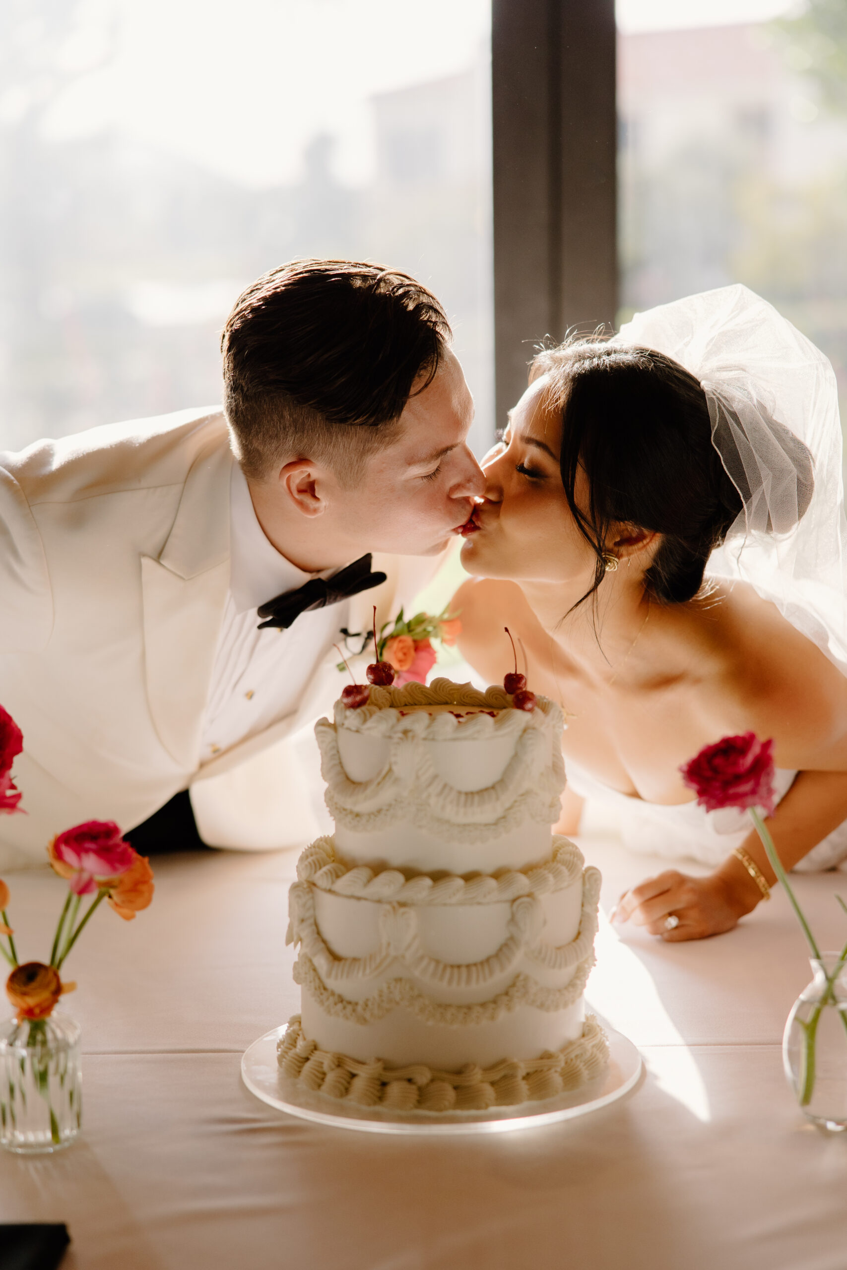 Bride and groom leaning in to kiss over their vintage-style wedding cake at McCormick Ranch Golf Club, sunlight glowing behind them.