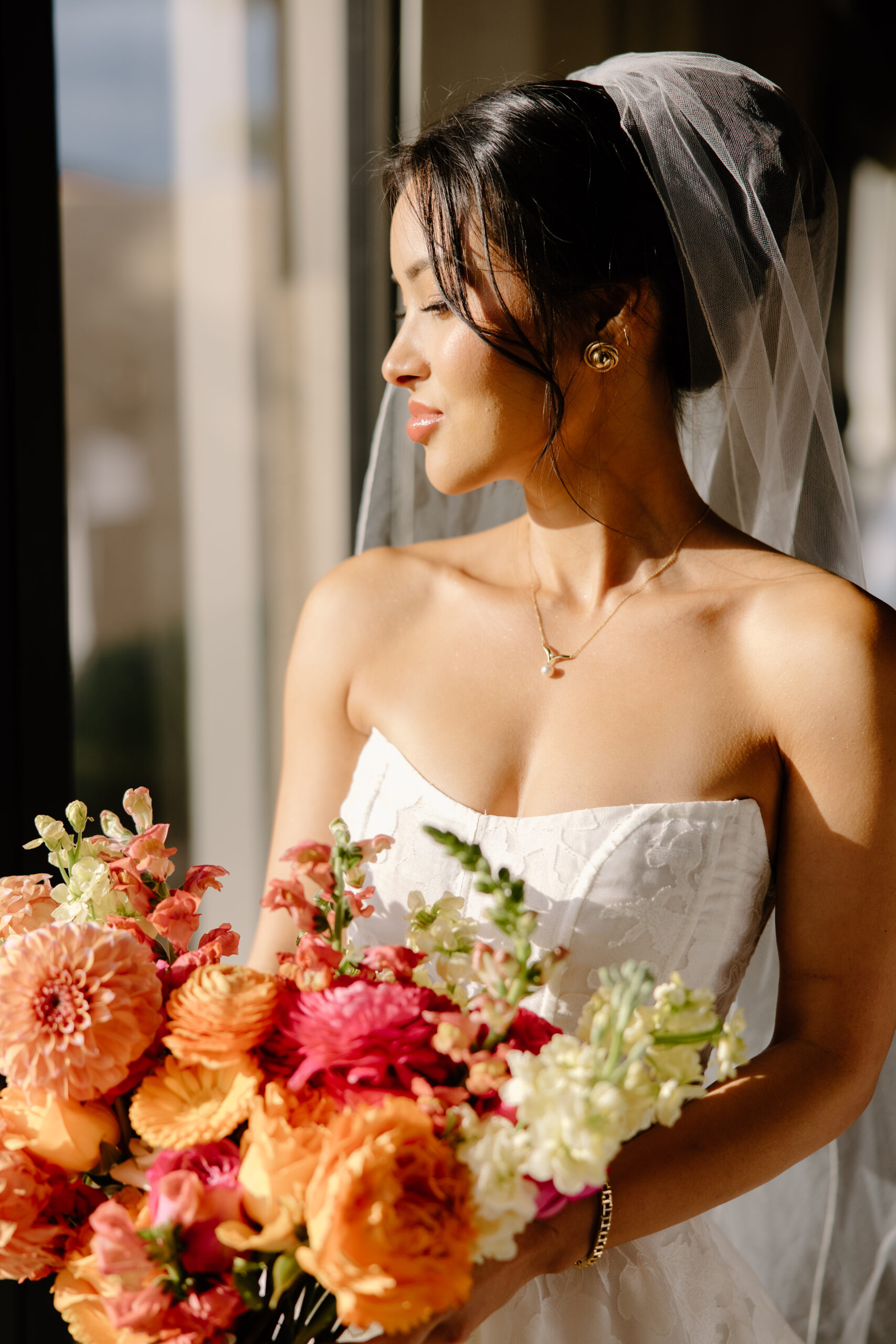 A bride standing by a sunlit window holding a vibrant bouquet of orange and pink flowers, her veil draped softly behind her.