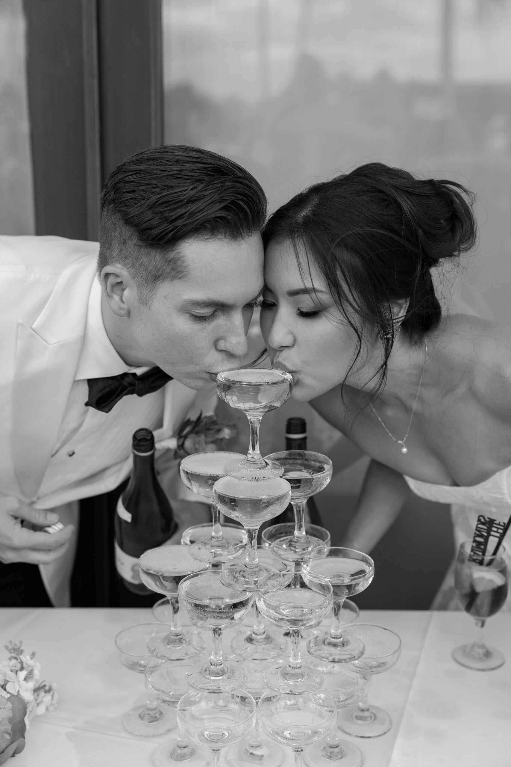 The bride and groom sipping from a champagne tower together in a playful black-and-white moment.