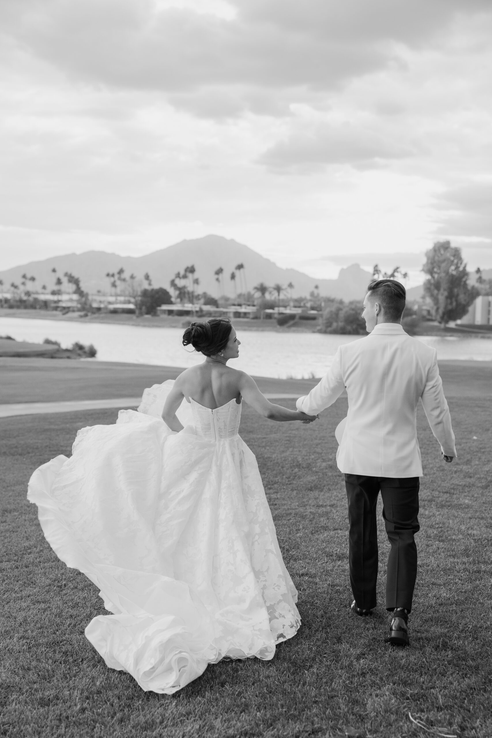 A bride and groom walking hand-in-hand across a grassy lawn with mountain views behind them at mccormick ranch golf club.