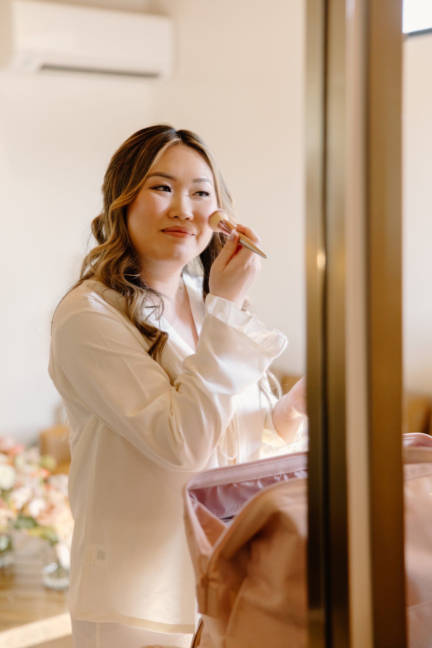 A bride getting her makeup applied in front of a mirror, her skin softly lit while a rose-gold brush is held near her face.
