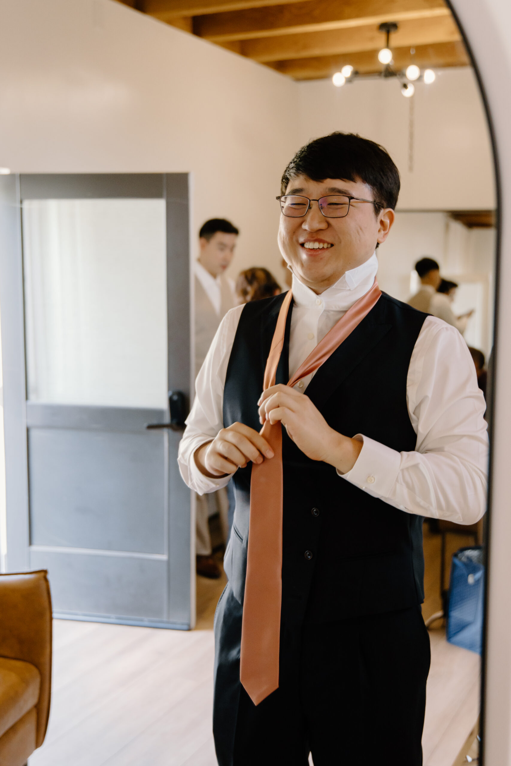 A groom smiling while adjusting his blush-colored tie, dressed in a white shirt and black vest inside a bright minimalist bridal suite at The Paseo, Arizona.
