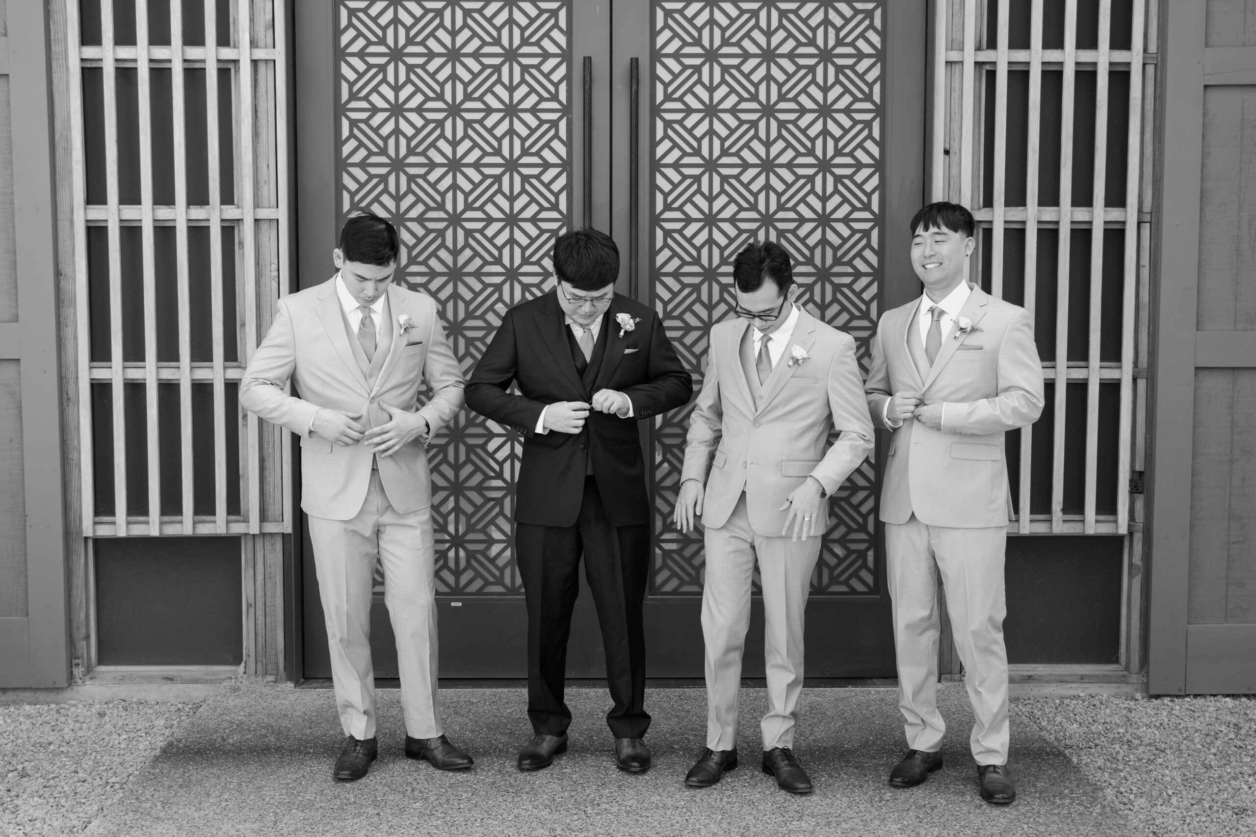 A black-and-white photo of four groomsmen in matching light-colored suits standing in front of geometric patterned doors, smiling and adjusting their jackets.