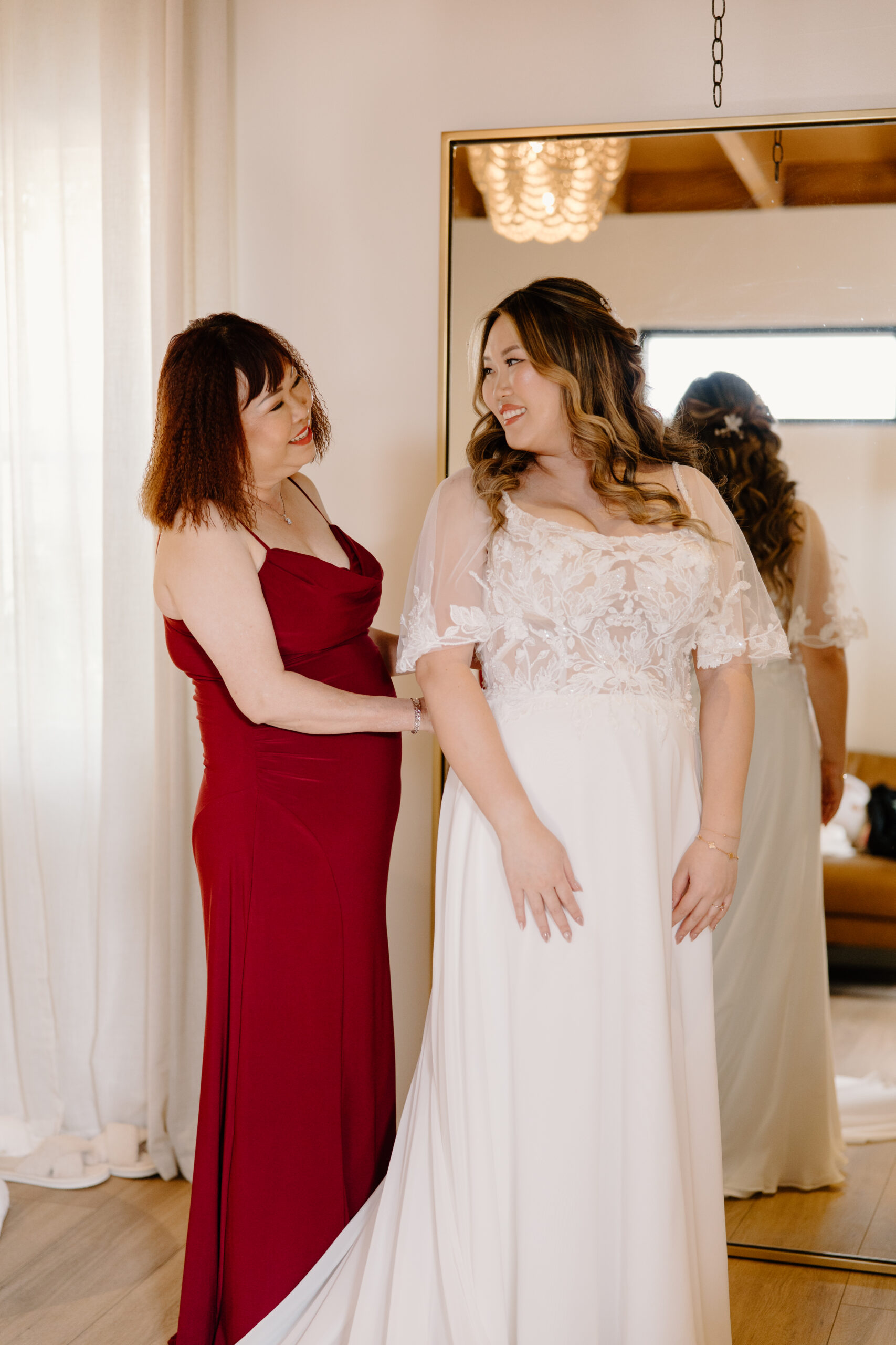 A mother helping the bride adjust her wedding dress, both smiling at each other inside a softly lit room with warm wood panels.