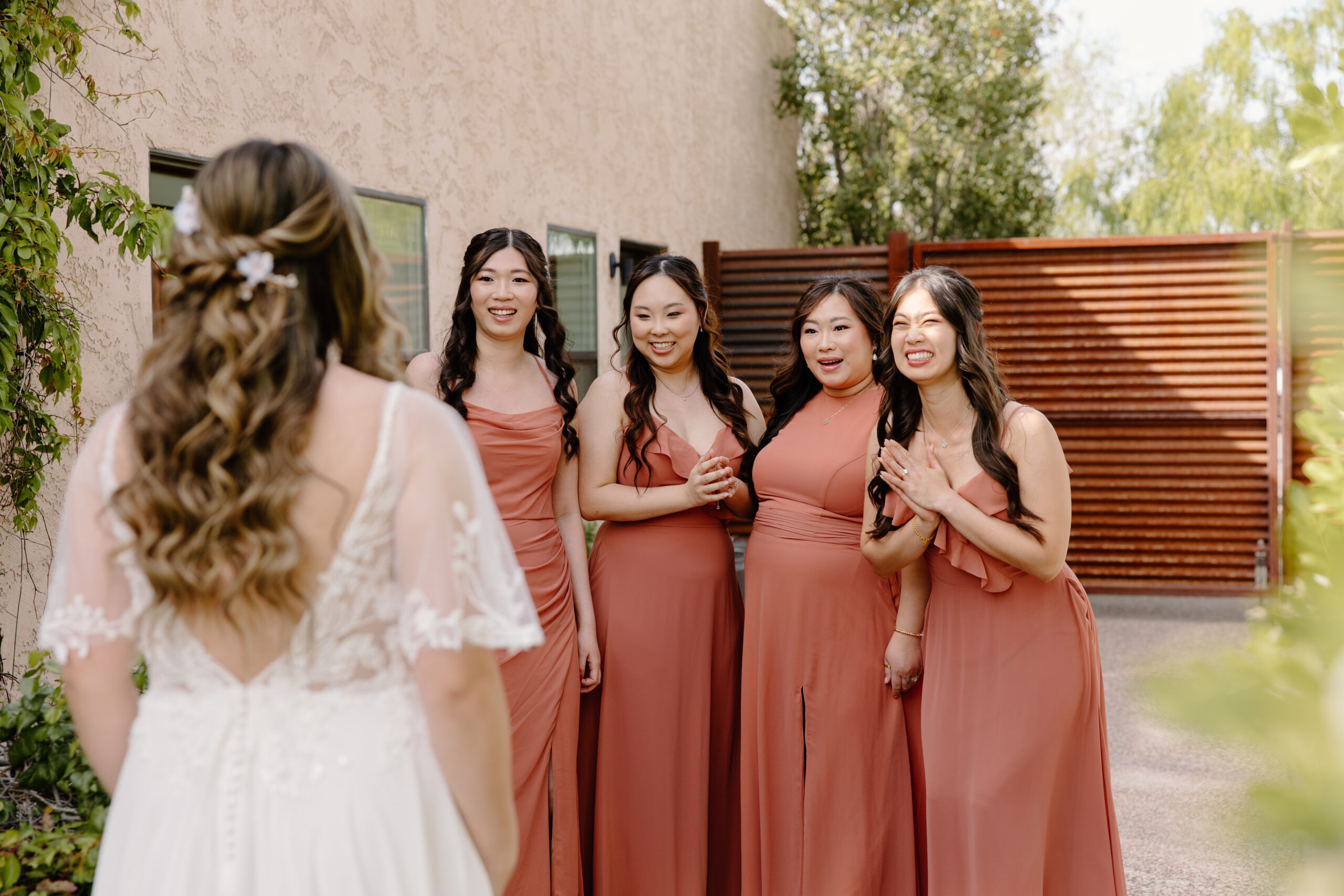 The bride doing a first look with her bridesmaids in a desert courtyard, all wearing terracotta-tone dresses and holding small bouquets, reacting excitedly.