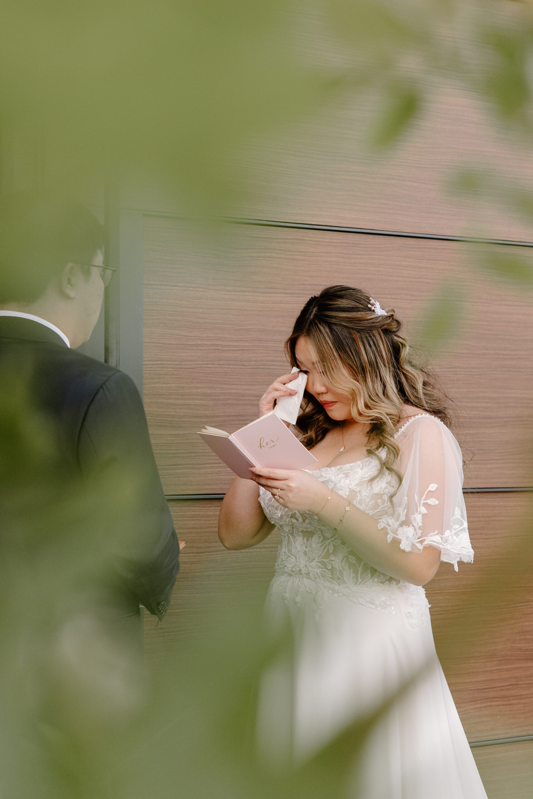 Bride reading handwritten vows from a small pink booklet, wiping a tear, while the groom smiles beside her in front of a modern wooden wall.