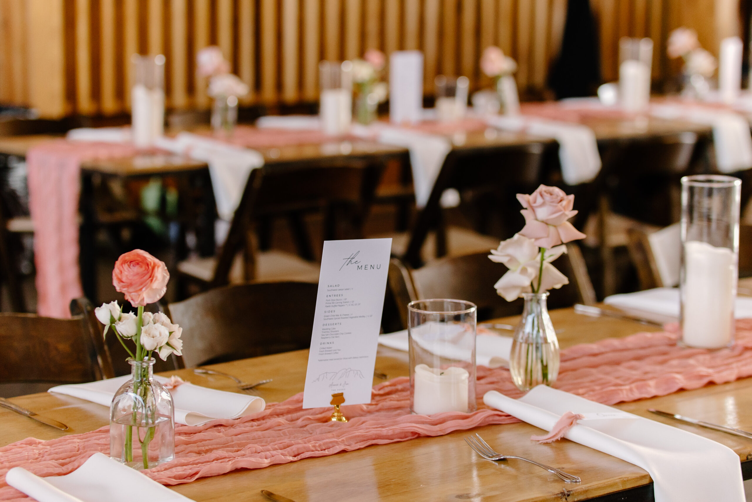 A detailed reception table setting with a textured runner, candles, and floral accents softly blurred behind it.
